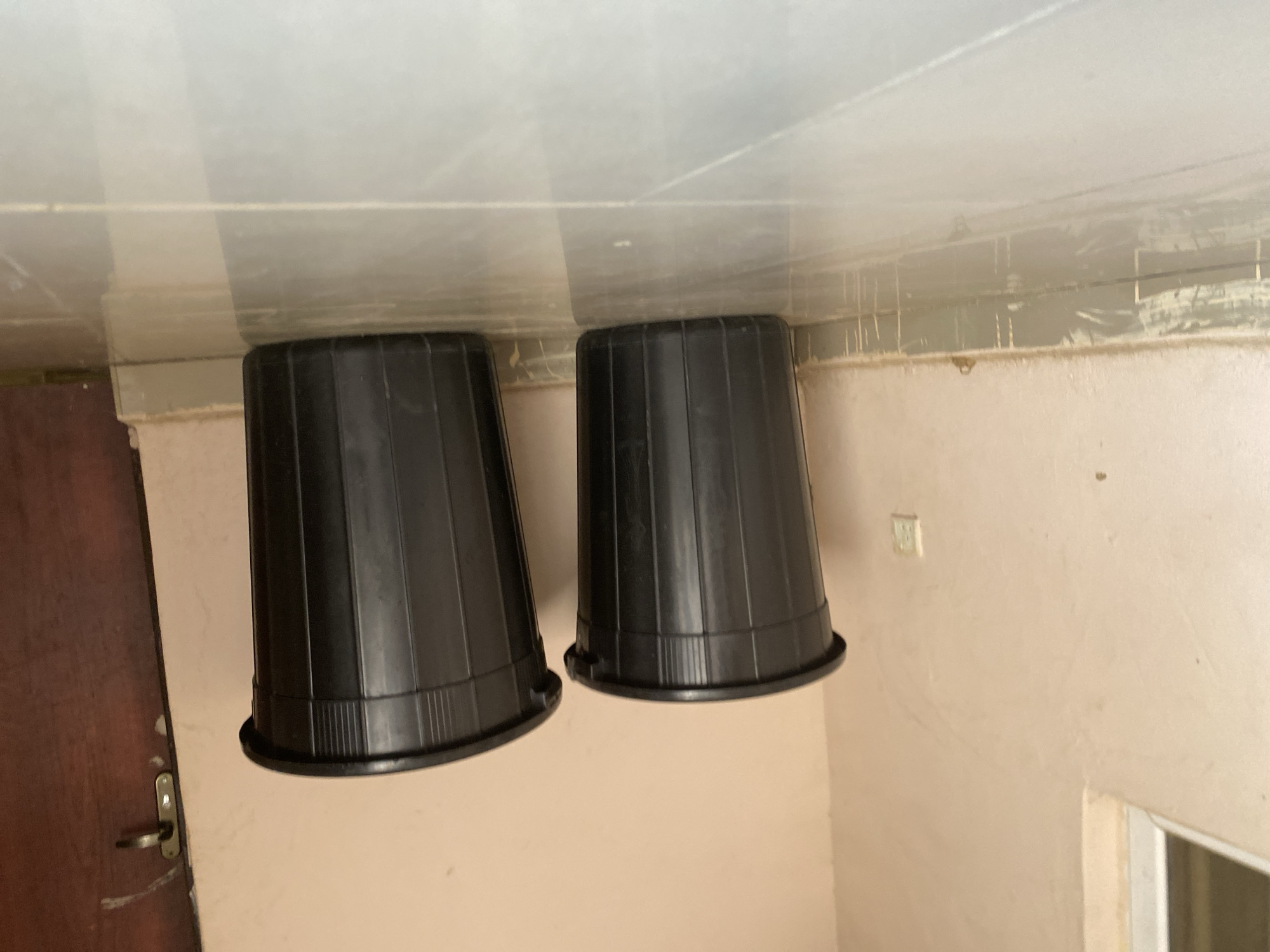 Two black plastic trash cans are positioned on a light-colored tiled floor in front of a cream-colored wall. The trash can on the left is slightly closer to the viewer, while the one on the right is further back and to the right. Both trash cans have a ribbed exterior and a wide rim. A dark brown door with a silver handle is visible to the far right of the frame, partially obscured by the trash can on the right. A window frame with a light-colored edge is visible in the upper left corner of the image. The base of the wall shows signs of wear, with a lighter colored border and some streaks or drips. The scene appears to be indoors, possibly in a hallway or room with a door and a window. The overall lighting is neutral.