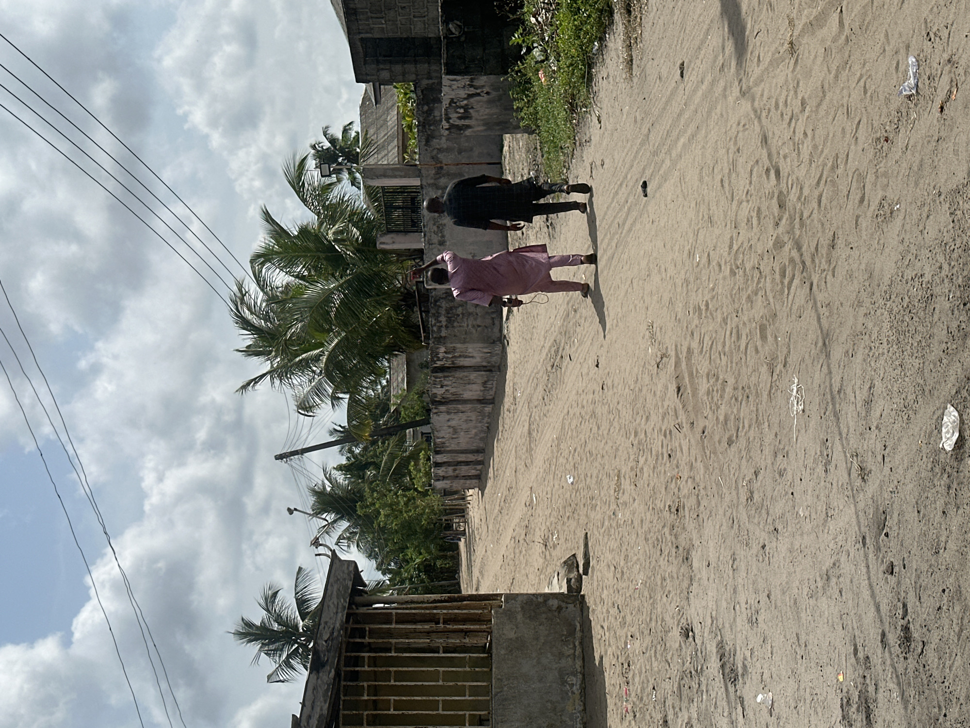 The image captures a daytime scene on an unpaved, sandy road in Badagry, Nigeria. Two men are walking away from the viewer along the path. The man in the foreground is dressed in a light pink traditional Nigerian outfit, possibly a kaftan or agbada, and appears to be holding a small object in his right hand. The man further ahead is wearing a dark-colored (likely black or navy) traditional outfit.
On the left side of the road, there are concrete walls and structures, some appearing weathered or unfinished, interspersed with lush green vegetation, including prominent palm trees. Overhead power lines crisscross against a partly cloudy sky. The ground is sandy with visible footprints and some scattered litter, including pieces of plastic. The right side of the road features a slightly raised dirt embankment with sparse weeds. The overall setting suggests a residential or semi-urban area with simple infrastructure, typical of a West African locale. There is no visible text in the image.