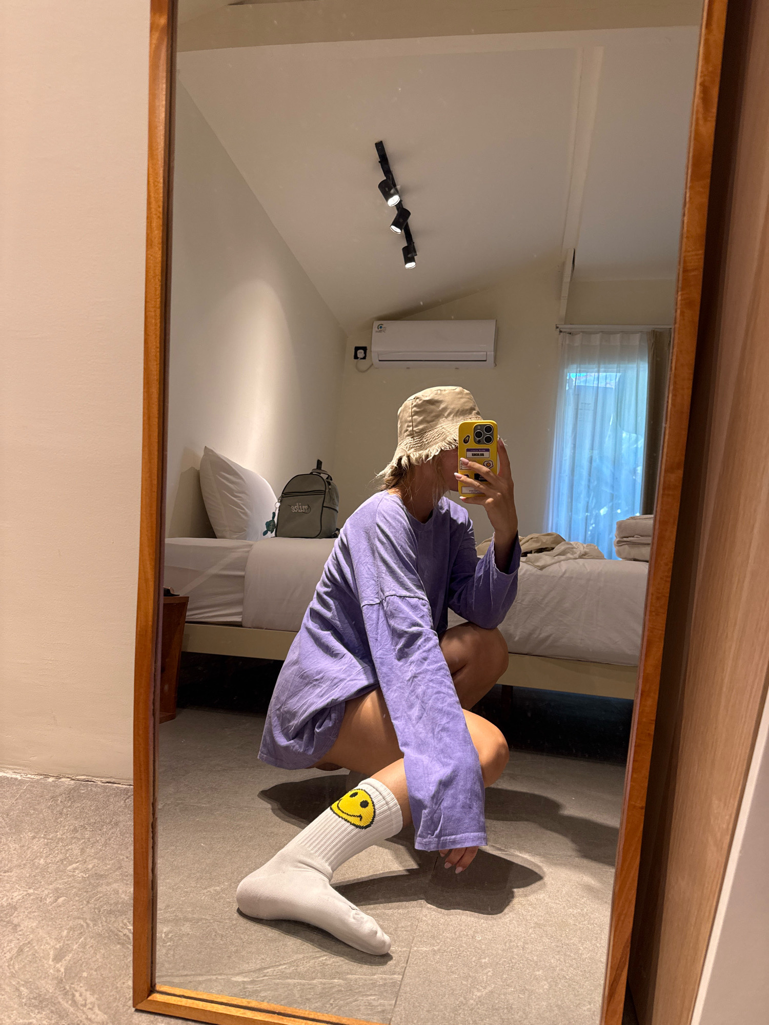 A young person is reflected in a large mirror, squatting on a gray tiled floor. They are wearing a purple long-sleeved shirt, shorts, a beige bucket hat, and white socks with a yellow smiley face design. The person is holding a yellow smartphone up to take the photo. In the reflection of the room behind them, there is a bed with white bedding, a white pillow, a gray backpack, a white air conditioner, and a track lighting fixture on the ceiling. A sheer white curtain covers a window to the right. The room appears to be a bedroom.