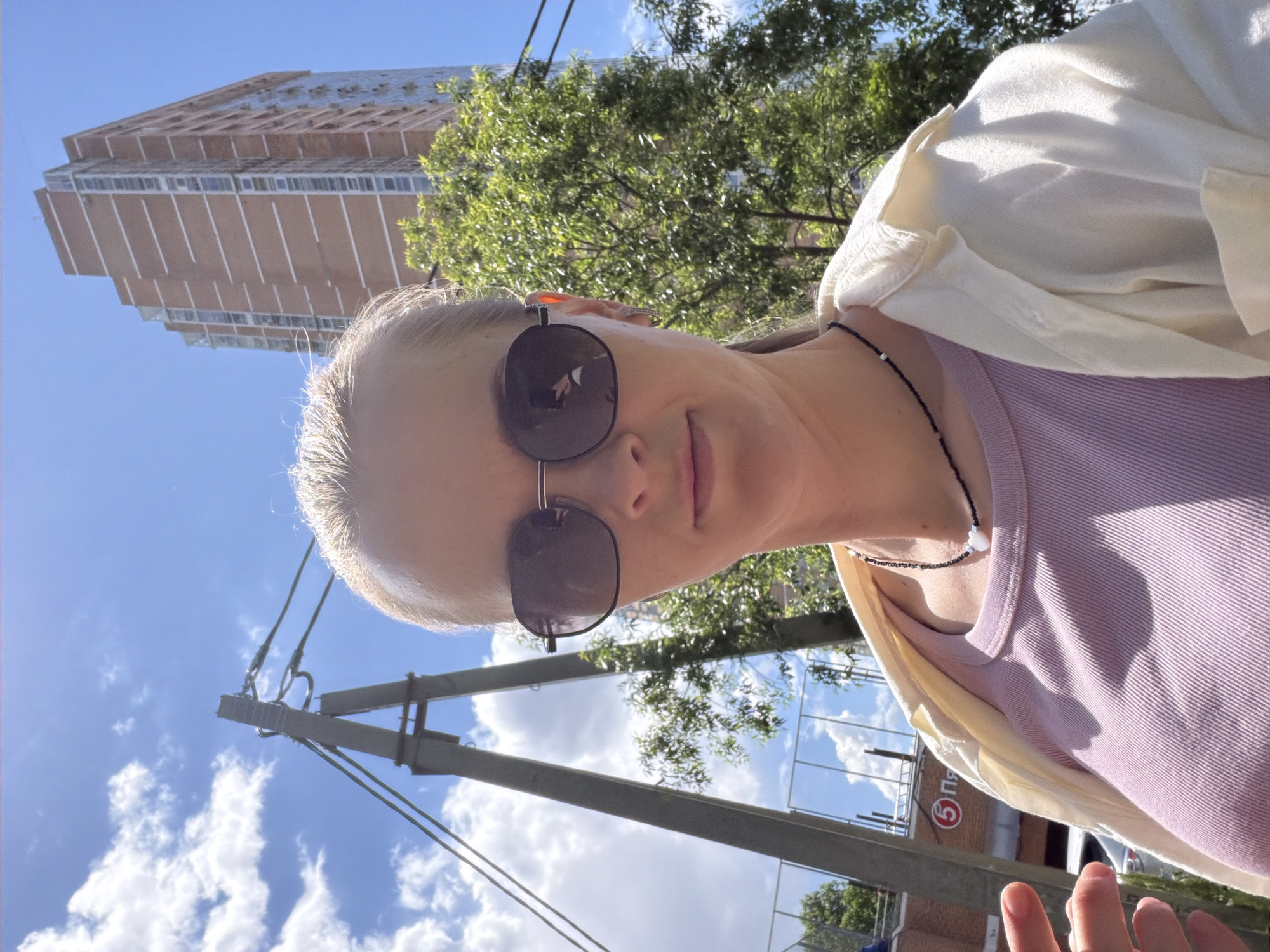 A young woman with blonde hair pulled back into a ponytail, wearing sunglasses, a light purple ribbed tank top, and an open cream-colored shirt is taking a selfie outdoors. She is wearing a necklace with black beads and a white heart pendant. Behind her, a tall brick building with many windows stands against a bright blue sky with scattered white clouds. To her left, a large grey metal structure with wires running between it and a pole is visible. Further to the left, a red sign with a white circled "5" and Cyrillic text is partially visible. The setting appears to be an urban environment, potentially near a transportation hub or public area in Khabarovsk, Russia, given the Cyrillic signage. Sunlight is strong, casting bright illumination and some shadows.