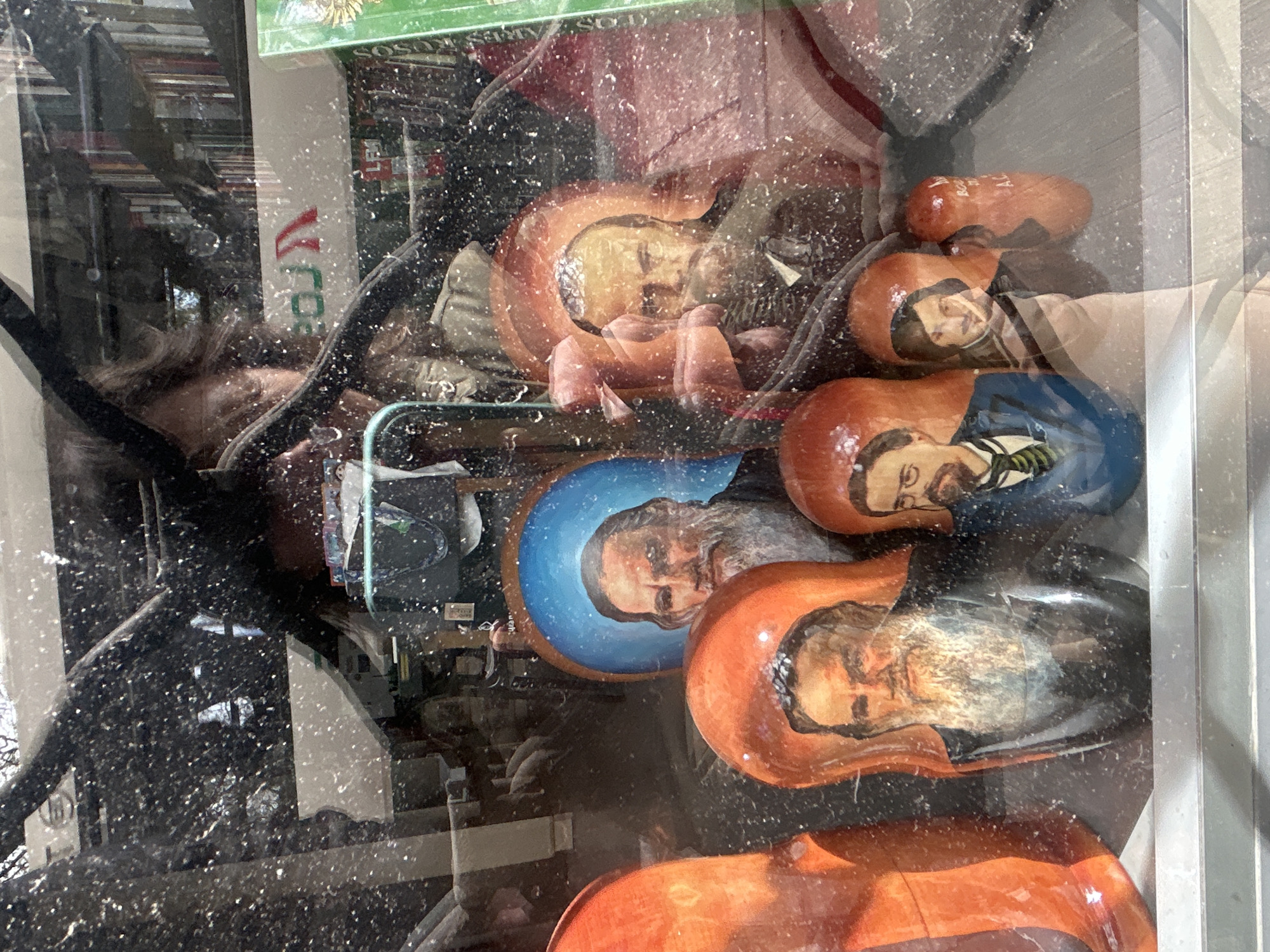 The image shows a display of matryoshka dolls, also known as Russian nesting dolls, through a glass window. The dolls are painted with portraits of famous figures. One doll depicts a man with a long white beard, wearing a dark garment and a blue halo. Another doll shows a man with a beard and glasses, wearing a suit and tie. A third doll has a man with a beard and a dark suit. A fourth doll shows a younger man with dark hair, wearing a light-colored garment. A fifth, smaller doll is partially visible. The dolls are arranged in a row, with some larger orange-colored dolls partially obscuring them.
The reflection in the glass shows a person holding a phone, possibly taking the photo, and bookshelves in the background. The scene is set in a shop, with signs visible in the reflection. One sign appears to read "LEO" and another has red and green lettering. Given the presence of "LEO" in the background, this location is likely within the El Ateneo Grand Splendid bookstore, a well-known landmark in the city of Buenos Aires, Argentina, famous for its architecture and extensive book collection. The overall impression is that of a cultural display within a bookstore.