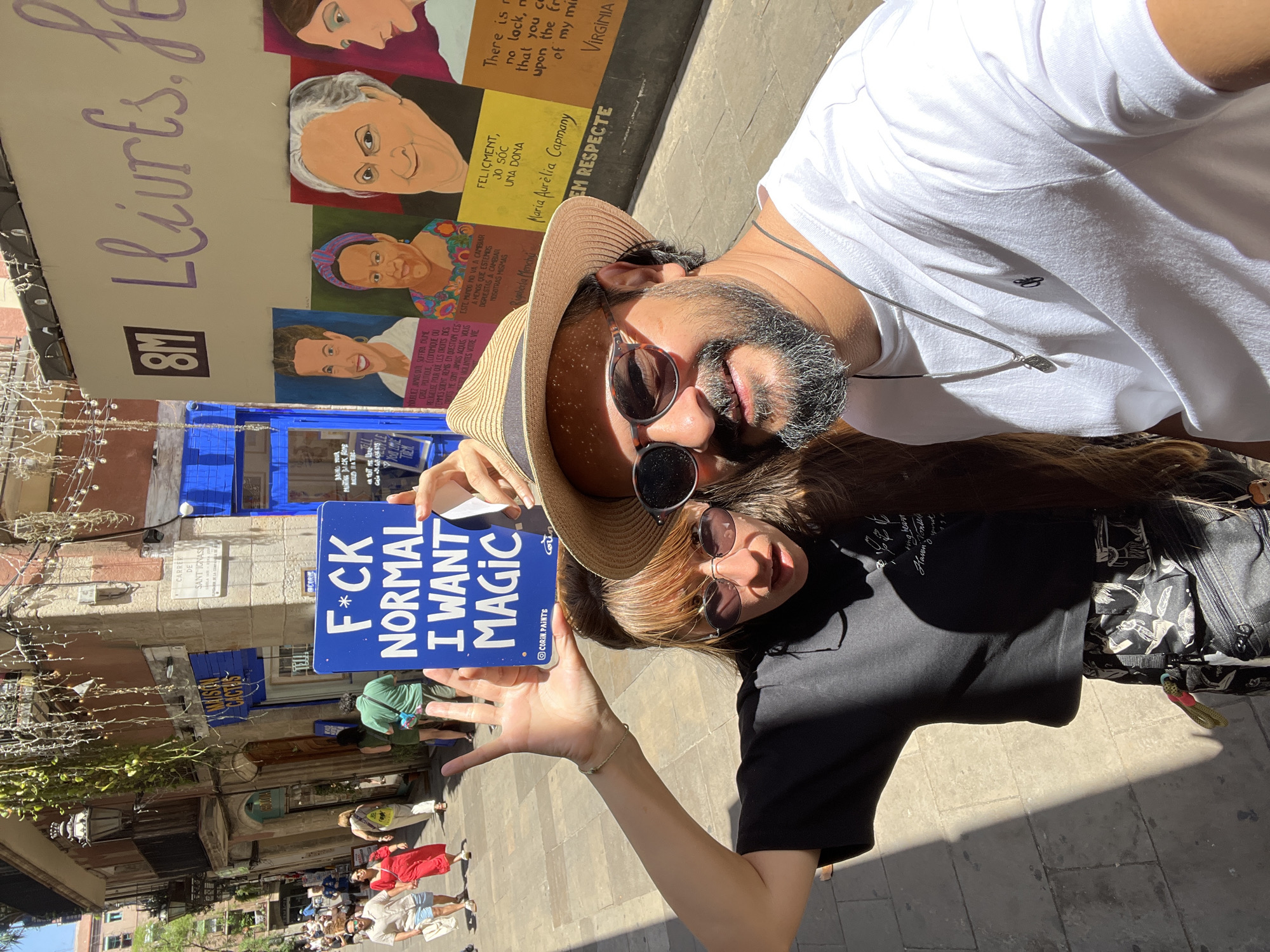 A couple poses for a selfie in a narrow street in Barcelona's Gothic Quarter. The woman on the left holds up a blue sign that reads "F*CK NORMAL I WANT MAGIC". She wears a black t-shirt, patterned shorts, sunglasses, and has long brown hair. The man on the right wears a white t-shirt, a straw fedora, and sunglasses. Behind them, a mural depicts four portraits of women with text in Catalan and Spanish, celebrating International Women's Day (8M). The buildings surrounding the street have traditional Barcelona architecture with balconies and stone facades. Sunlight casts shadows on the cobblestone street, indicating a bright, sunny day. A store named "MAISON CACTUS" is visible on the left.