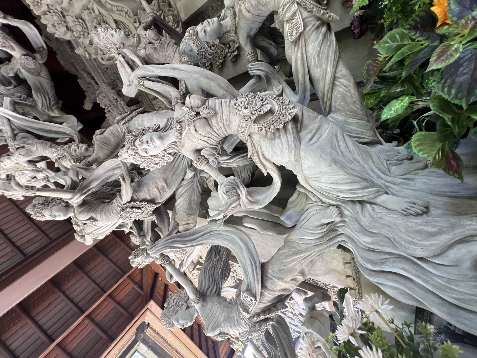 The image depicts a section of a Balinese architectural element, specifically a sculpted frieze. The frieze is comprised of numerous figures, predominantly female, rendered in a light-colored material such as stone or plaster. These figures are intricately detailed with ornate headdresses, flowing garments, and expressive faces. The arrangement suggests a dynamic composition, with figures intertwined and gesturing.
The location is likely Kedewatan, Indonesia, based on the Balinese style of the sculpture. There are wooden beams and window frames that provide structural context to the sculpture. Some foliage with white and purple flowers and green leaves are situated near the right side of the frieze, adding a natural element to the man-made artwork.