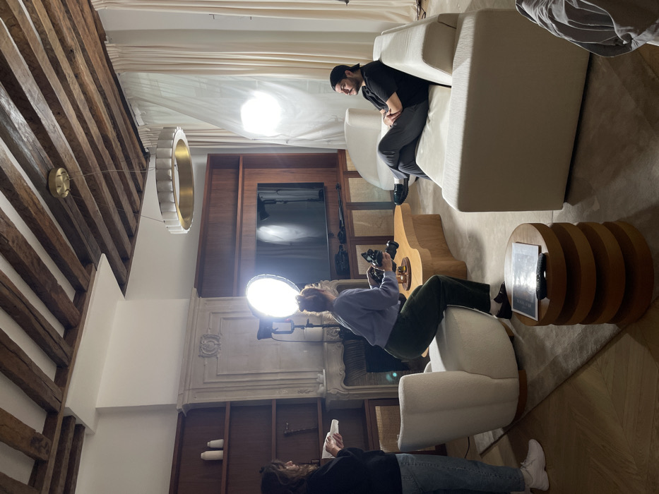 The image depicts an indoor photoshoot taking place in a renovated apartment within a historic Parisian building. The room features a high ceiling with prominent exposed dark wooden beams, white walls, and dark wooden built-in shelving units. An ornate white marble fireplace mantel is visible on the left, with a large flat-screen television integrated into the wooden shelving unit. Light-colored sheer curtains cover windows on the right, indicative of a large window opening.
Three individuals are present. On the right, a male subject with dark hair and a beard, wearing a backward black baseball cap, black t-shirt, and dark pants, is seated with crossed arms on a white modular sofa, looking towards the left. Centrally, a female photographer with reddish-brown hair tied in a bun, wearing a light purple long-sleeved top and dark green trousers, is squatting by a light wood, organic-shaped coffee table, holding a black DSLR camera aimed at the male subject. On the far left, another female with dark wavy hair, dressed in a dark top and light jeans, stands holding a white bottle or dispenser.
The scene is actively lit by two large, circular studio lights equipped with softboxes, one positioned behind the photographer and another on the right side, illuminating the male subject. A modern, circular gold-toned chandelier hangs from the ceiling. The floor consists of light wood parquet, partially covered by a large light grey area rug. Additional furniture includes a white curved upholstered armchair, two other white armchairs in the background, and a stacked cylindrical wooden side table holding a magazine and remote controls. The individuals are engaged in producing photographic or video content.
