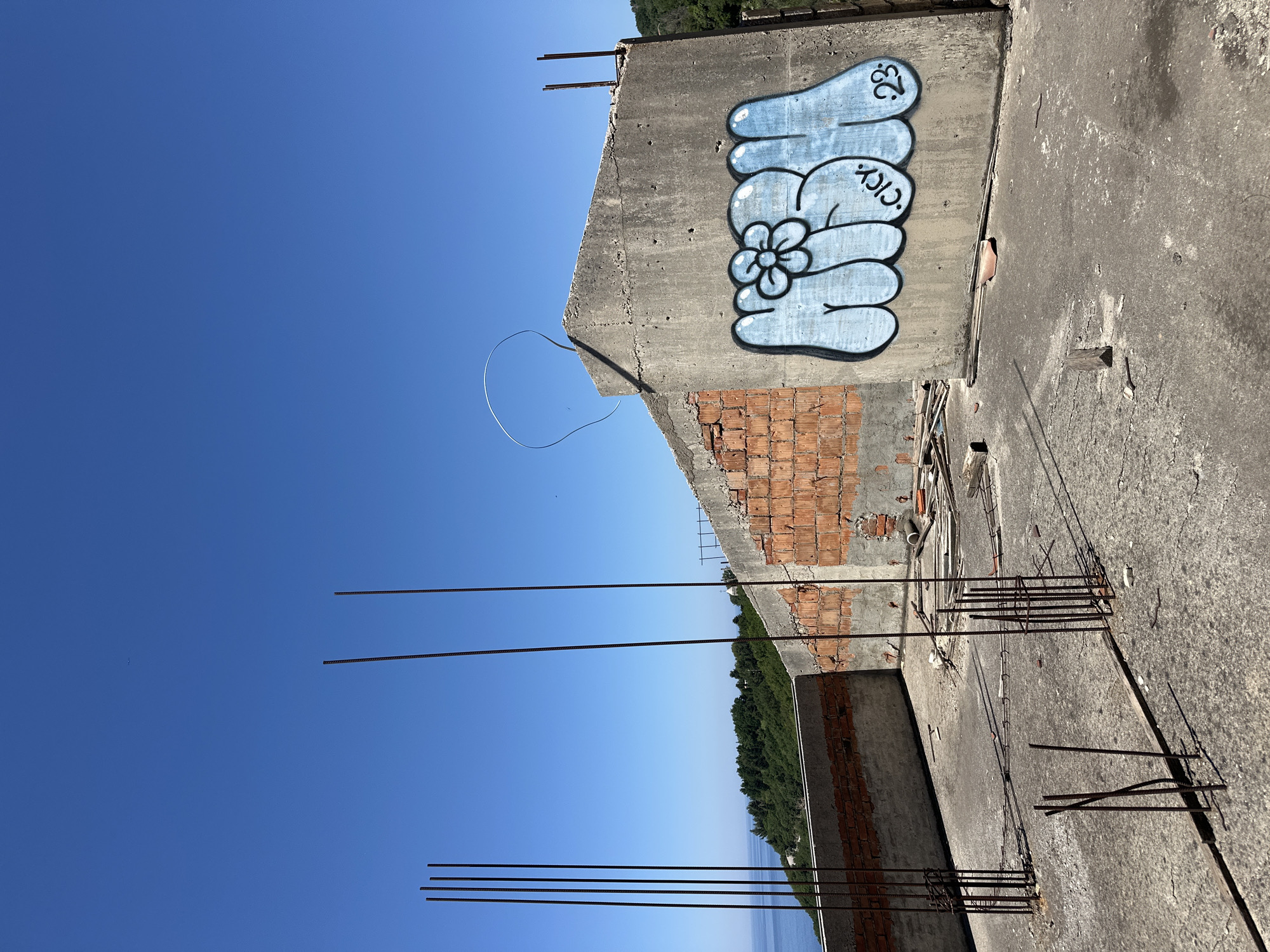 The image shows a building under construction in Gornji Katun, Montenegro. The walls are a mix of concrete and brick. Graffiti art is visible on the concrete wall. Rebar extends upward from the concrete platform, suggesting the structure is incomplete. The sky is a clear blue. In the background, there are trees and a glimpse of the sea.