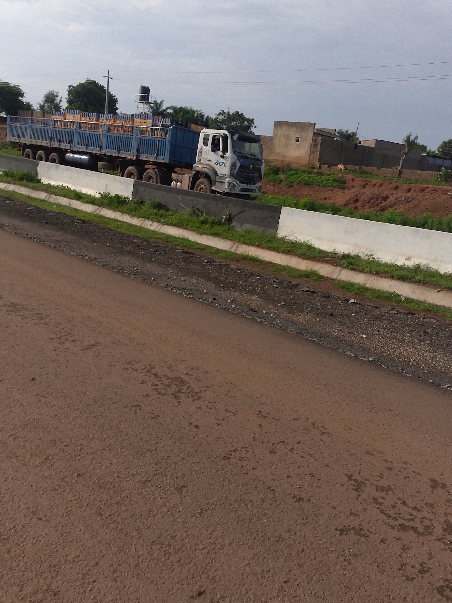 A long, blue flatbed truck is parked on the side of a paved road. The truck is laden with stacked wooden crates, mostly covered by a blue tarp. The truck has a white cab with "GPC" logo on the side. The truck has multiple axles and a fuel tank visible underneath. The road appears to be a new construction, with a dirt shoulder and a concrete barrier. In the background, there are some buildings under construction, trees, and a water tower. The sky is overcast. The scene is likely in Keffi, Nigeria, as suggested by the presence of a water tower commonly seen in Nigerian towns, and the general architecture of the buildings under construction.