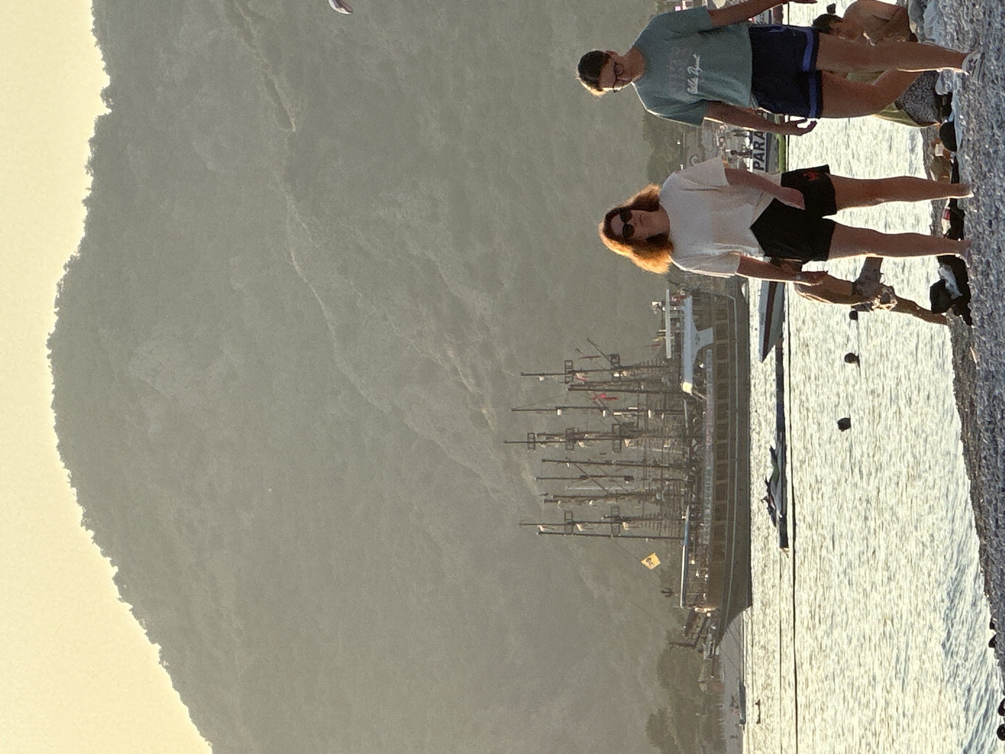 This image captures a coastal scene in Fethiye, Turkey, at what appears to be late afternoon or early evening. The foreground features a pebbled beach, where two individuals are walking from right to left. The person on the left is a female with reddish-brown, backlit hair, wearing sunglasses, a white t-shirt, and black shorts, walking barefoot. The person on the right is a female with dark hair, wearing eyeglasses, a light teal t-shirt, dark shorts, and light-colored shoes.
In the mid-ground, a large, multi-masted wooden boat resembling a historical sailing ship or a tour boat is positioned on the water. It displays a yellow flag on one of its masts and other smaller flags. Several smaller white motorboats and a jet ski are also visible on the water, with a black buoy floating to the right. To the left of the walking individuals, a man is partially submerged in the water, and further left, two heads indicate other people swimming. On the beach to the right of the walkers, indistinct figures are visible, including a man sitting with his back to the camera. A white and red striped parasail is partially visible in the upper right portion of the sky, indicating airborne activity.
The background consists of large, hazy mountains covered in sparse vegetation, appearing somewhat muted by the atmospheric conditions. The sky above the mountains is a clear, light yellow-orange, reflecting the time of day. The water surface is calm and reflects the warm light, creating a shimmering effect.