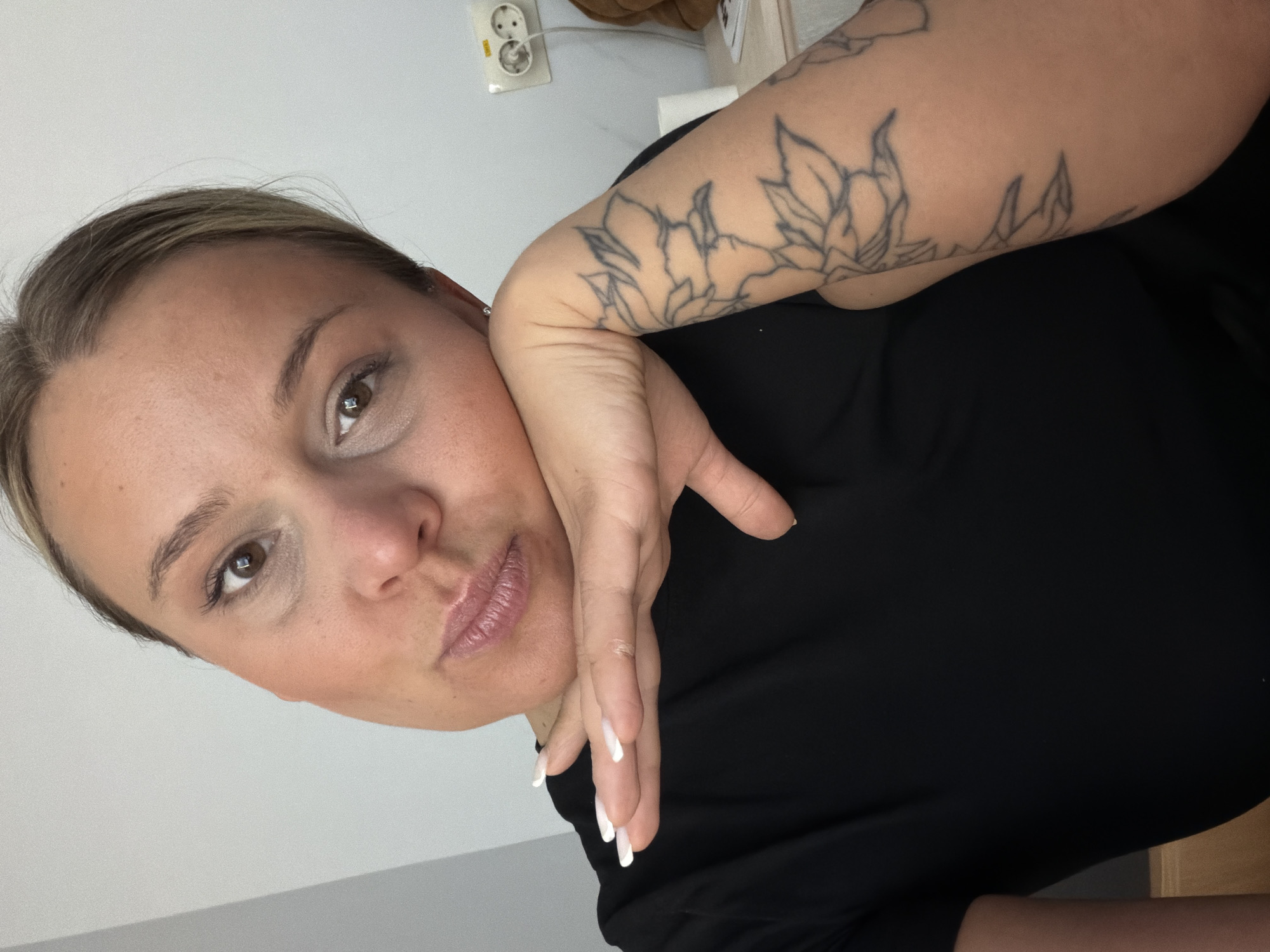 A woman is posing for a photo. She has fair skin, light brown hair pulled back, and brown eyes. She is wearing a black shirt and has a floral tattoo on her right arm. Her nails are painted white. She is holding her hand to her face, with her cheek resting on her fingers.
In the background, there is a white wall with a European-style electrical outlet. Based on the outlet type, it is plausible that this image was taken in the city of Sarapul, Russia.