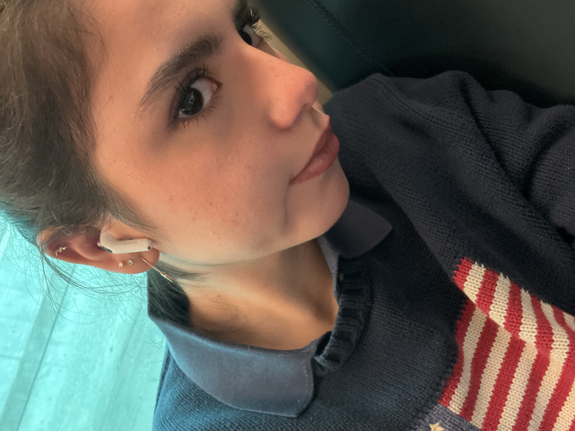A young woman is pictured from a low, close-up angle. She has dark hair pulled back, fair skin, and is wearing an earbud in her left ear. She is looking upwards with a slight expression. She's wearing a navy blue polo shirt with a sweater over it that has an American flag design on the chest. The background is blurred, suggesting she might be near a window or a light-colored surface. The location cannot be determined to be Chicago, United States of America from the details of the image.
