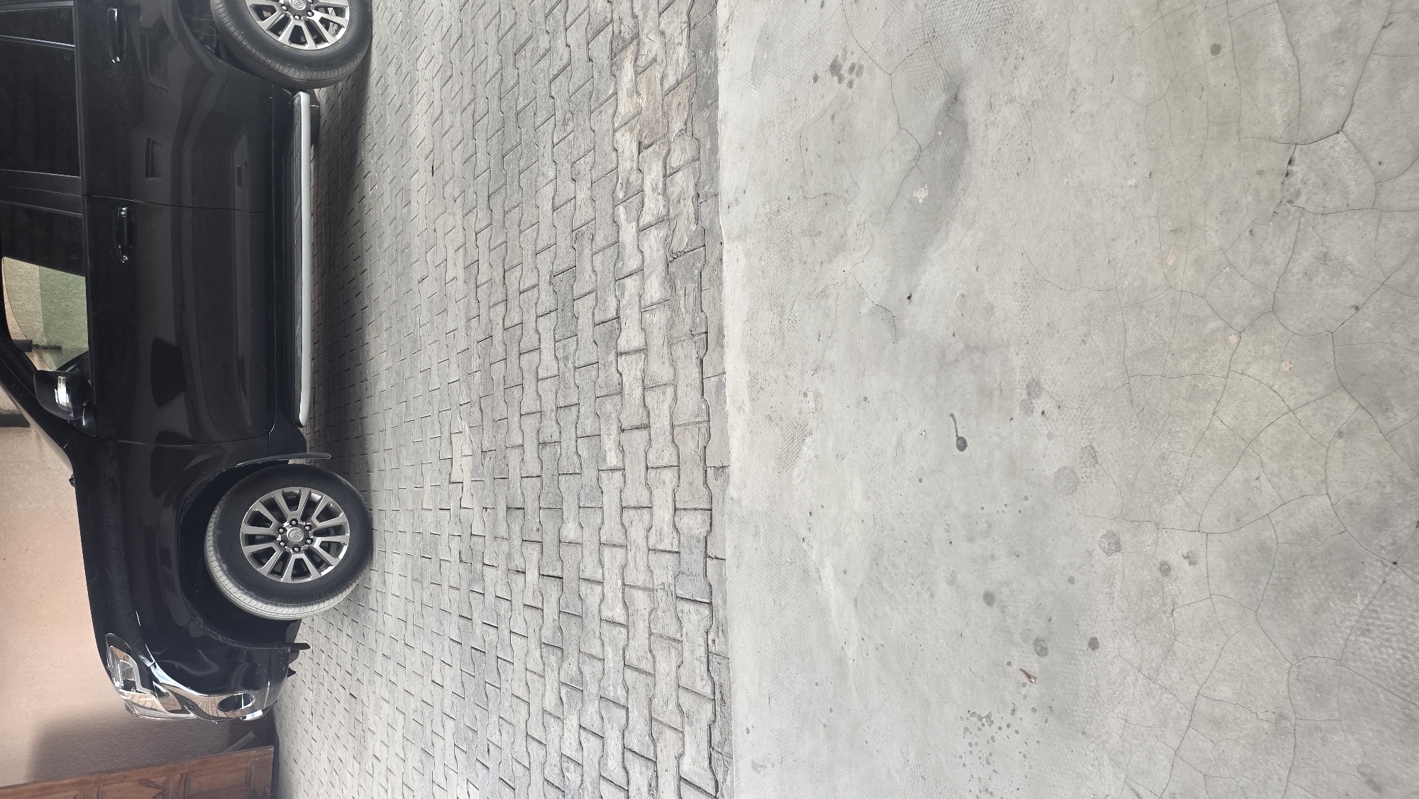 A black SUV is parked on a patterned concrete driveway in Chuy, Kyrgyzstan. The vehicle is facing right, and only the front half of the car is visible. The SUV has alloy wheels and a silver running board. The driveway is made of interlocking concrete pavers. The ground in front of the pavers is a rough, cracked concrete surface. There is no visible human activity in the image.
