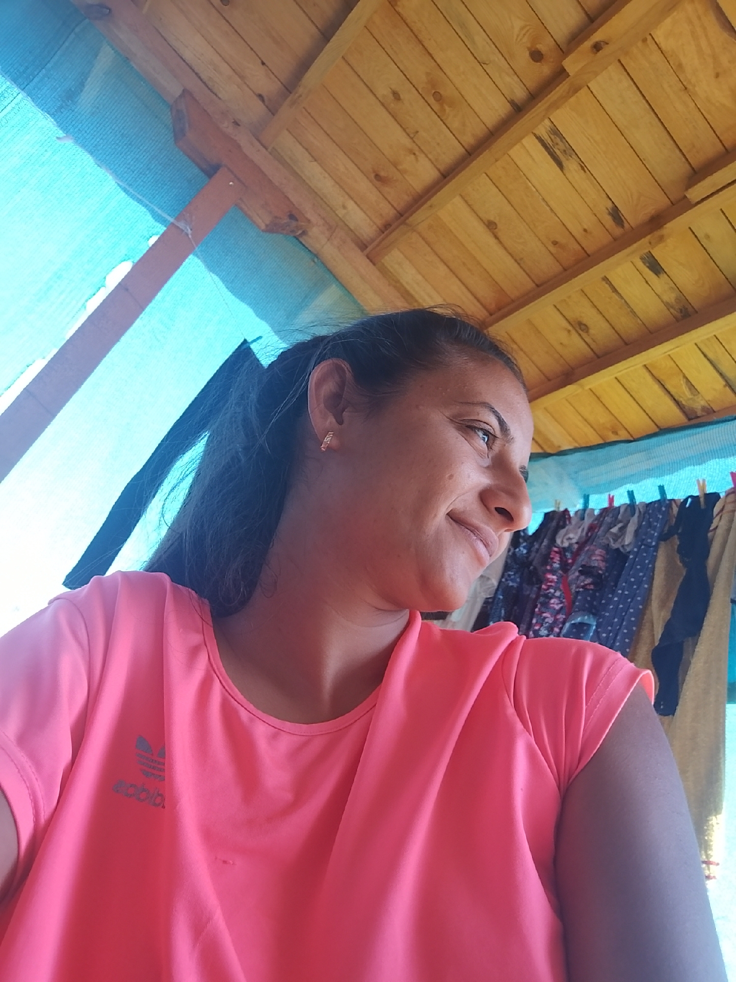 A medium shot shows a woman from the chest up, looking to her right, outdoors under a wooden roof structure covered by a blue tarp. She has dark hair pulled back into a ponytail and is wearing a bright pink t-shirt with a grey Adidas logo on the left chest. She has a gold hoop earring in her visible ear. Behind her, to the right, clothes hang on a clothesline. The wooden beams and planks of the roof are visible above her. This scene is located in Zimnitsa, Bulgaria.