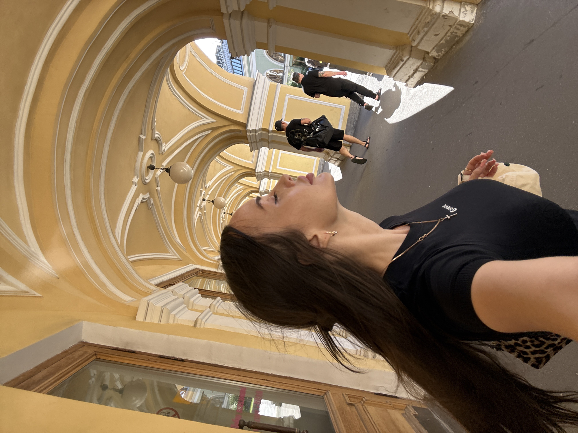 In Saint Petersburg, Russia, a woman with long dark hair, wearing a black t-shirt and a necklace with a cross pendant, is seen from the side, sticking her tongue out. She is walking in an arched, yellow-painted passageway with white trim, which appears to be a public area, possibly part of a historic building or colonnade. Sunlight streams into the passageway, casting shadows on the paved ground. Two men are walking away from the camera in the distance, one carrying a black bag. The architectural style suggests a European, likely Russian, urban setting.