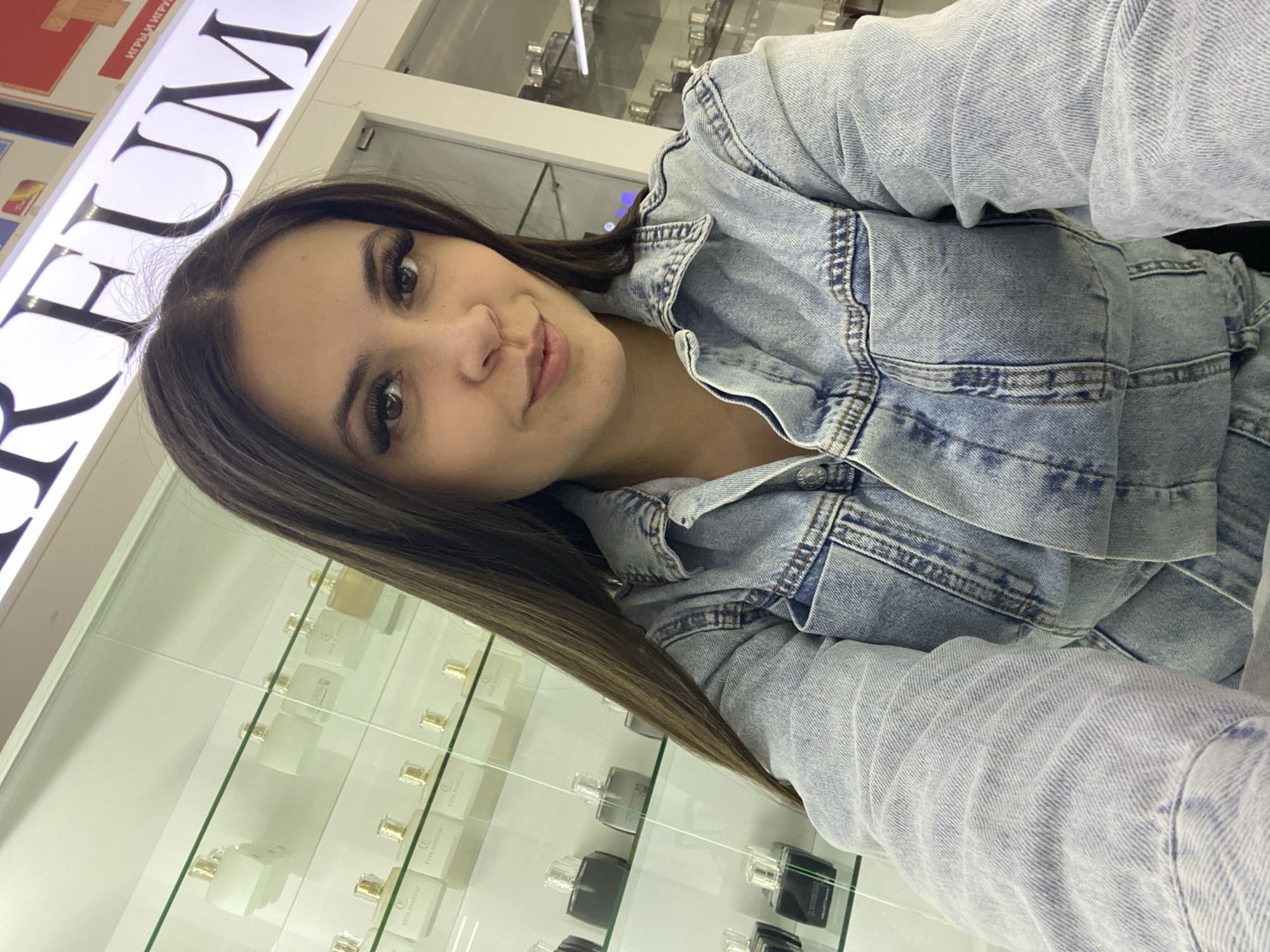 The image is a selfie taken by a young woman inside a retail establishment, likely a perfume or cosmetics store, located in Chaikovsky, Russia. The woman, with long dark hair and dark eyes, occupies the left side of the frame, looking towards the camera with a subtle, content smile. She is wearing a light blue, faded denim jacket, which is visible across her shoulders and arms.
In the background, an illuminated white sign with large, dark capital letters displays part of a word, "FUM," which is likely "PERFUM" or "PARFUM." Above this, partially obscured red signage shows Cyrillic text, which appears to be "ИГРЫ И ИГРУ" (Games and Game) or similar, indicating other products or departments nearby. Below and to the right of the woman, clear glass display cases are filled with various perfume bottles. Many of these bottles are small, with some appearing in clear, black, or gold-capped designs. Text like "ETID PERFUM" is visible on some of the bottles. The store's interior is brightly lit, creating a clean and modern ambiance. The image is captured at an angle, characteristic of a casual selfie.