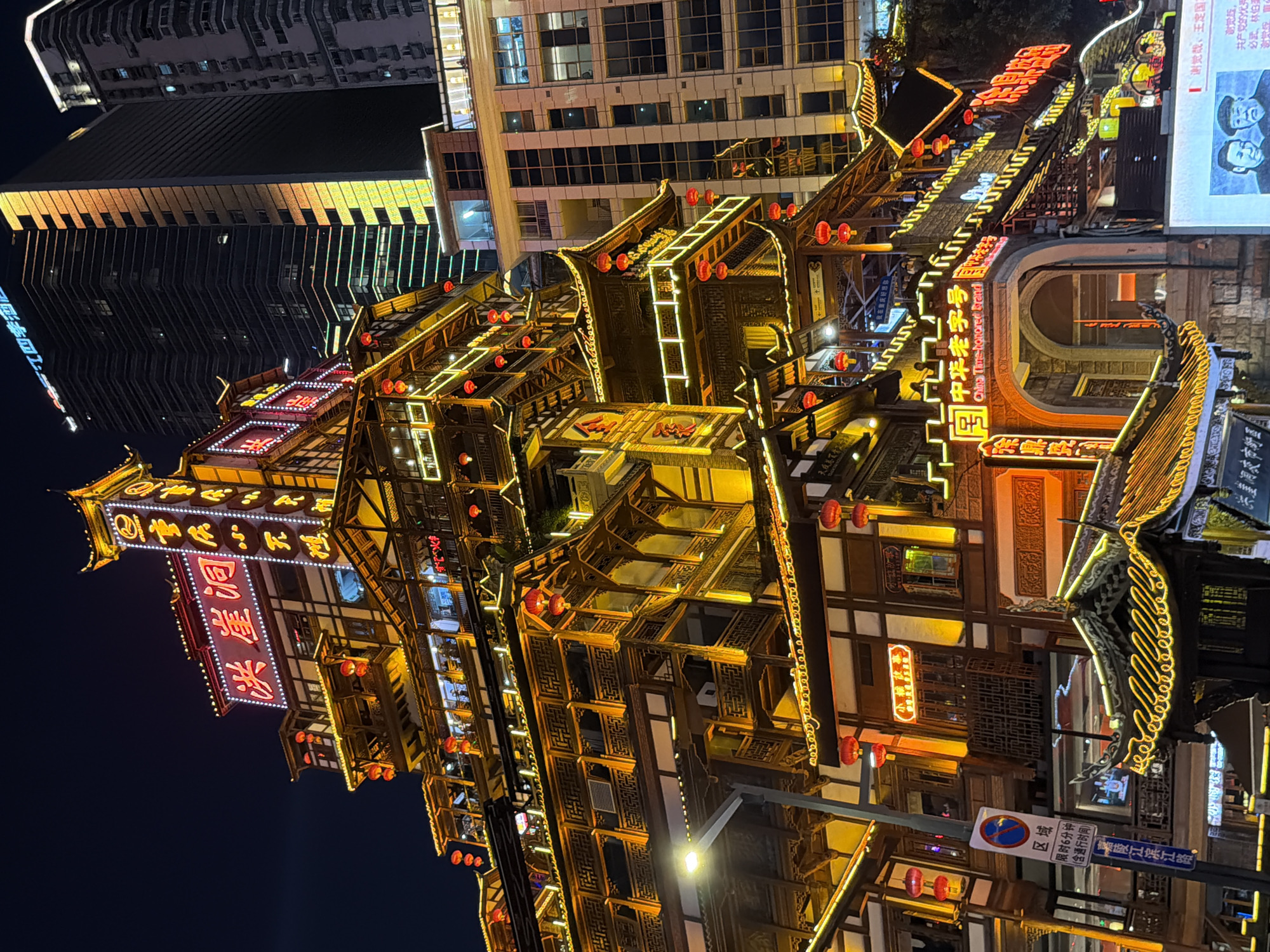 This media file captures a nighttime scene in the Yuzhong District of China, likely at the popular Hongyadong complex. The primary subject is a multi-story traditional Chinese building adorned with bright neon lights and red lanterns. The architecture is intricate, featuring wooden elements, patterned windows, and tiered roofs. The building is illuminated with a warm yellow and white glow from the extensive lighting, creating a vibrant and festive atmosphere.
The surrounding environment includes modern high-rise buildings in the background, contrasting with the traditional structure in the foreground. A streetlamp is visible, indicating it's a public area. Signs with Chinese text are present, including one that reads "中华老字号" (China Time-honored Brand) and another that appears to be directional signage with traffic regulations, mentioning a limited parking time of 6 minutes.
The image is characterized by the interplay of light and shadow, with the neon signs casting a glow on the building's facade and the night sky. There are no people clearly visible in the frame. The overall impression is one of a bustling, illuminated urban landscape at night, showcasing a blend of traditional and modern Chinese architecture. The time of day is clearly night due to the darkness and the prominent artificial lighting. The weather appears to be clear, as there are no indications of rain or fog.