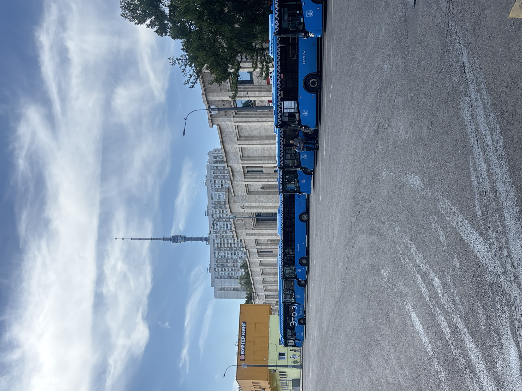 The image shows a row of blue public transport buses parked in a lot in front of a long, tan building. The building appears to be a commercial establishment with the Burger King logo visible on a smaller orange building attached to the left side of the tan structure. Behind the buildings, a tall, slender radio tower, identifiable as the Ostankino Tower, stands prominently against a cloudy blue sky. Residential buildings are also visible in the background, suggesting an urban environment. Several people are seen walking near the buses and entering/exiting the tan building. The ground in the foreground is asphalt with some white markings. The overall scene depicts a daytime view of a public transportation hub or depot in Moscow, Russia.