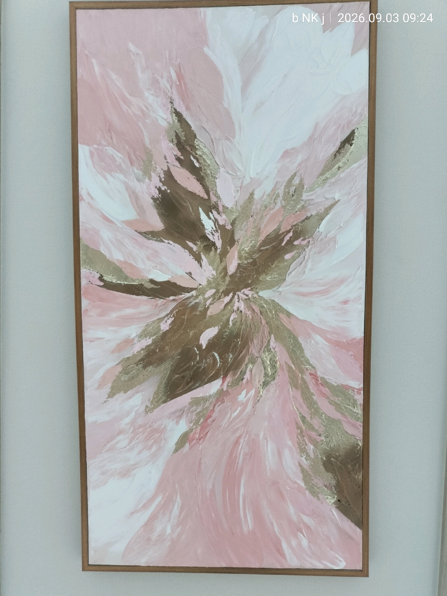 A vertical, framed abstract painting is displayed on a plain, light-colored wall. The painting features a textured, impasto style with broad brushstrokes. The dominant colors are various shades of pink and white, interspersed with metallic gold and brown accents. The composition radiates outwards from a central point, creating a dynamic, almost floral or feathery effect. The frame is a simple, light-colored wood. In the upper right corner of the image, a timestamp overlay reads "b NK j | 2026.09.03 09:24". There are no people or other objects visible in the immediate vicinity of the painting. The environment appears to be an interior space with neutral lighting and a simple wall. The location is identified as South Jakarta, Indonesia.