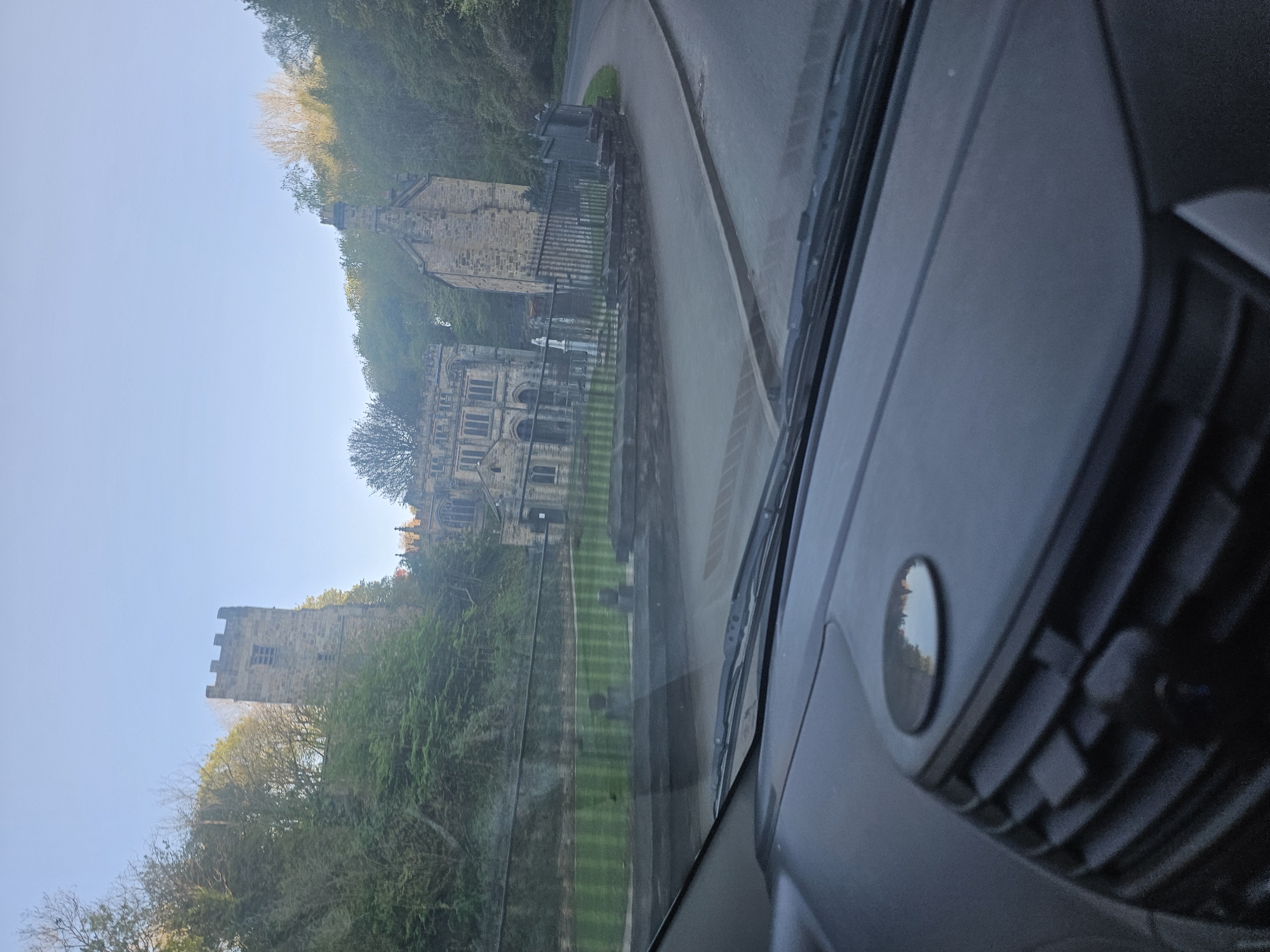 The image presents a picturesque view of a historic landscape, captured from the interior of a vehicle. The lower part of the frame reveals the dark grey dashboard and a portion of the windshield, including an air vent and a circular control, indicating the perspective of a passenger or driver.
In the mid-ground, a dark tarmac road, appearing damp or wet, curves gently from the left towards the right. Alongside the road, a neatly mown green lawn with distinct stripes extends towards a series of ancient stone buildings.
Two prominent historical structures dominate the scene. On the right, a large, multi-story stone building features numerous arched windows and intricate stonework, resembling an old church, abbey, or grand estate. To its left and slightly further back, a sturdy, square stone tower with battlements and a single visible window stands, partially framed by trees. The architectural style suggests a significant historical presence, characteristic of the United Kingdom.
Beyond the buildings and rising up a gentle incline, a dense canopy of green trees covers the hillside, with some deciduous trees appearing to have lighter or sparser foliage, possibly indicating early spring or late autumn. Above the trees and buildings, a clear, bright blue sky completes the tranquil setting.
The image captures a static moment during daylight hours, likely morning or late afternoon, given the clear sky and even lighting. The wet road, juxtaposed with the clear sky, suggests recent rain has passed, leaving the landscape refreshed. No specific activity is taking place other than the observation of the scenic view from the vehicle. There is no visible text within the image.