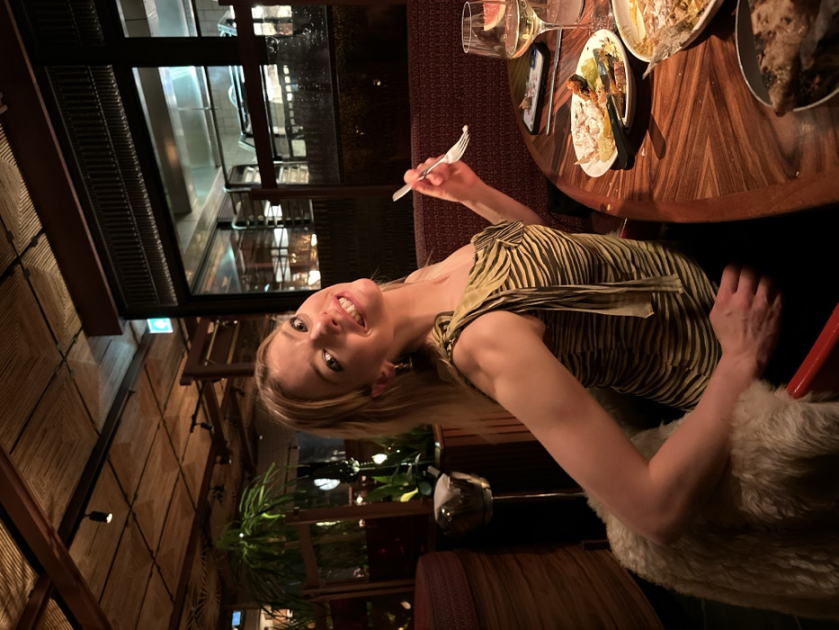 An indoor, eye-level shot captures a young woman smiling at the camera while dining at a restaurant in London, United Kingdom. She is seated at a round, dark wooden table with a distinctive patterned surface, holding a silver fork in her right hand. The woman has blonde hair, light skin, and is wearing a striking olive green and cream striped top, reminiscent of a zebra pattern. Her expression is bright and cheerful, with a wide smile directed towards the viewer.
On the table in front of her are several small plates, some containing food remnants, including what appears to be a skewer and various small dishes, suggesting a tapas or shared-plate dining experience. A wine glass filled with a light beverage and garnished with a slice of citrus (likely grapefruit) sits near a dark-colored smartphone. Another clear glass is also visible. The woman is seated on a chair or banquette adorned with a soft, light-colored, possibly fur or sheepskin, cushion or throw.
The background reveals a sophisticated restaurant interior. To the left, a textured wall with woven, square-patterned panels extends upwards. Dark wooden beams and structural elements are visible throughout the space. Large windows in the background look out onto an illuminated internal area, possibly a kitchen or another part of the establishment, suggesting it is evening or night outside. Lush green plants add a touch of nature to the refined setting. No visible text is discernible in the image. The overall atmosphere appears warm, inviting, and social.