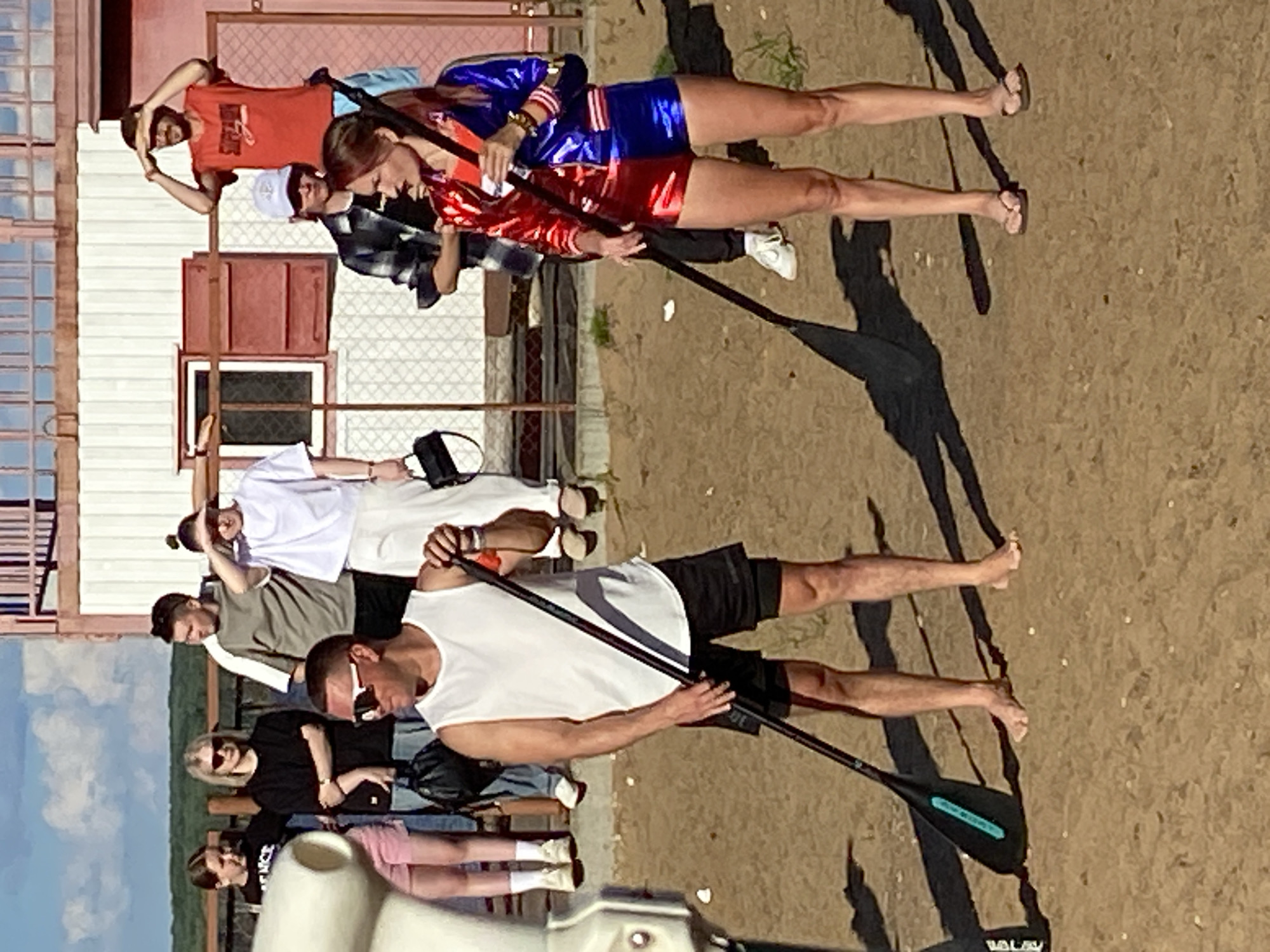 A group of people are standing on a sandy area near a wooden structure in Chaikovsky, Russia. In the foreground, a man wearing a white tank top and black shorts holds a paddle. To his right, a woman in a red and blue jacket and shorts also holds a paddle. Behind them, several other individuals are visible, some standing on a raised wooden platform, and others standing on the sand. The environment appears to be outdoors with a clear sky.