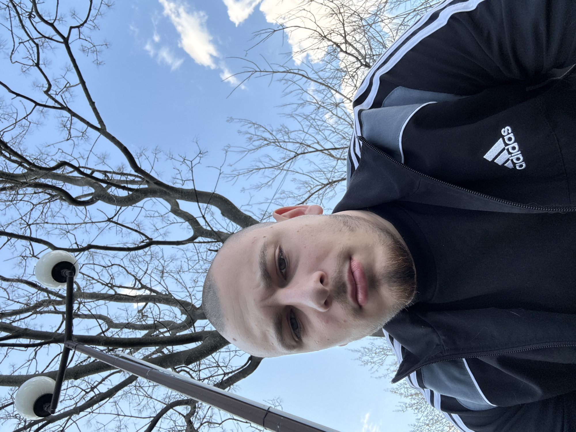 A low-angle selfie of a man outdoors, with a tree and a lamp post in the background, taken in Stara Zagora, Bulgaria.
The man has short, light-colored hair and a short beard. He is wearing a black jacket with white stripes on the sleeves and an "adidas" logo on the right side of the jacket. The sky is light blue with scattered white clouds visible through the branches of a leafless tree. In the lower-left corner, there is a lamp post with two globe-shaped lamps. The image is rotated counter-clockwise.