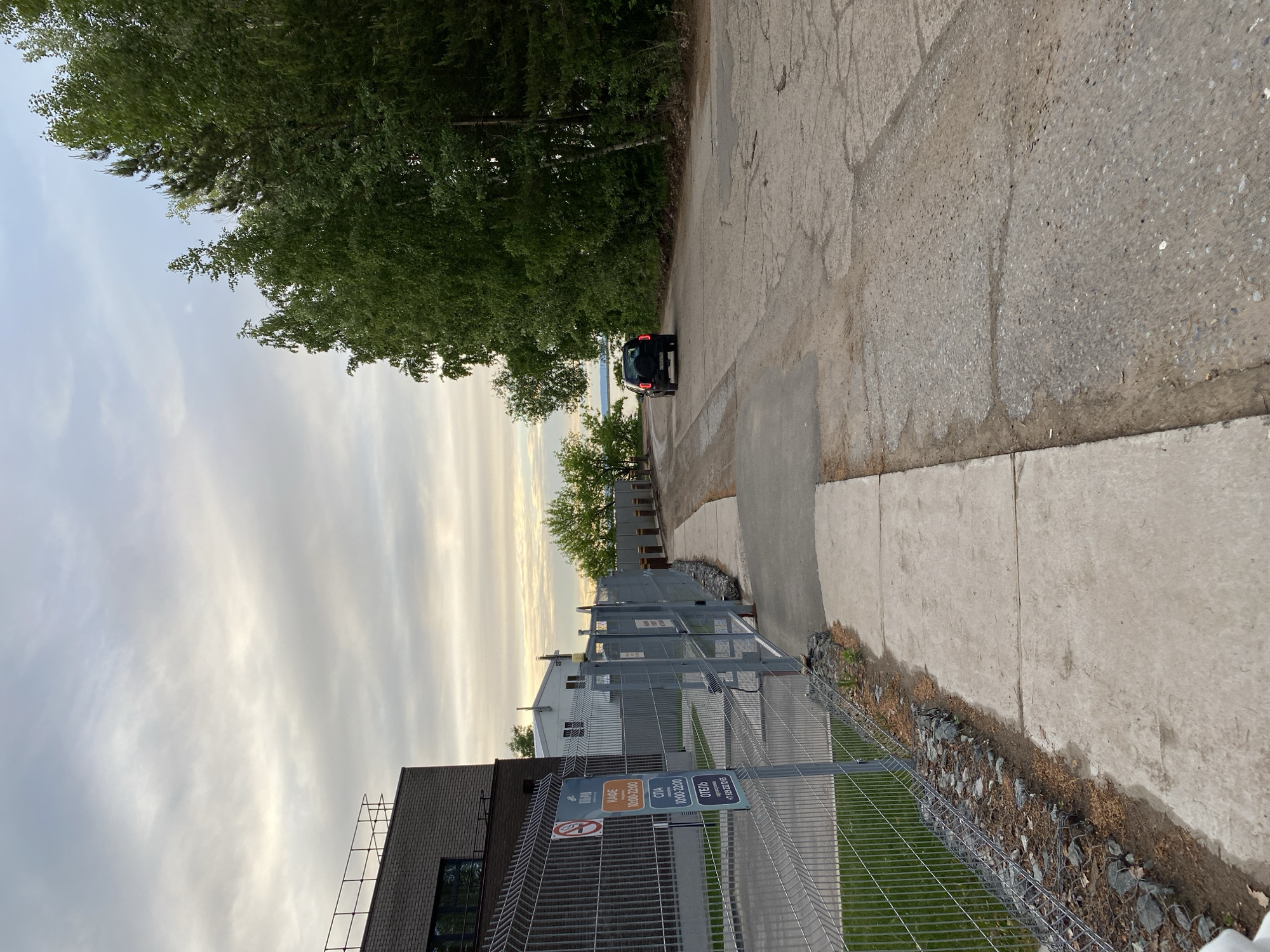 The image depicts a street scene in Chaikovsky, Russia. The photograph is taken at an angle, with the road receding into the distance. A black SUV is parked near the end of the road. To the left of the road is a grassy area enclosed by a wire fence. Adjacent to the fence is a building with a sign indicating the presence of a café (КАФЕ), spa (СПА), and hotel (ОТЕЛЬ), with operating hours and a phone number displayed. The sky is overcast with streaks of clouds. Trees line the left side of the road.