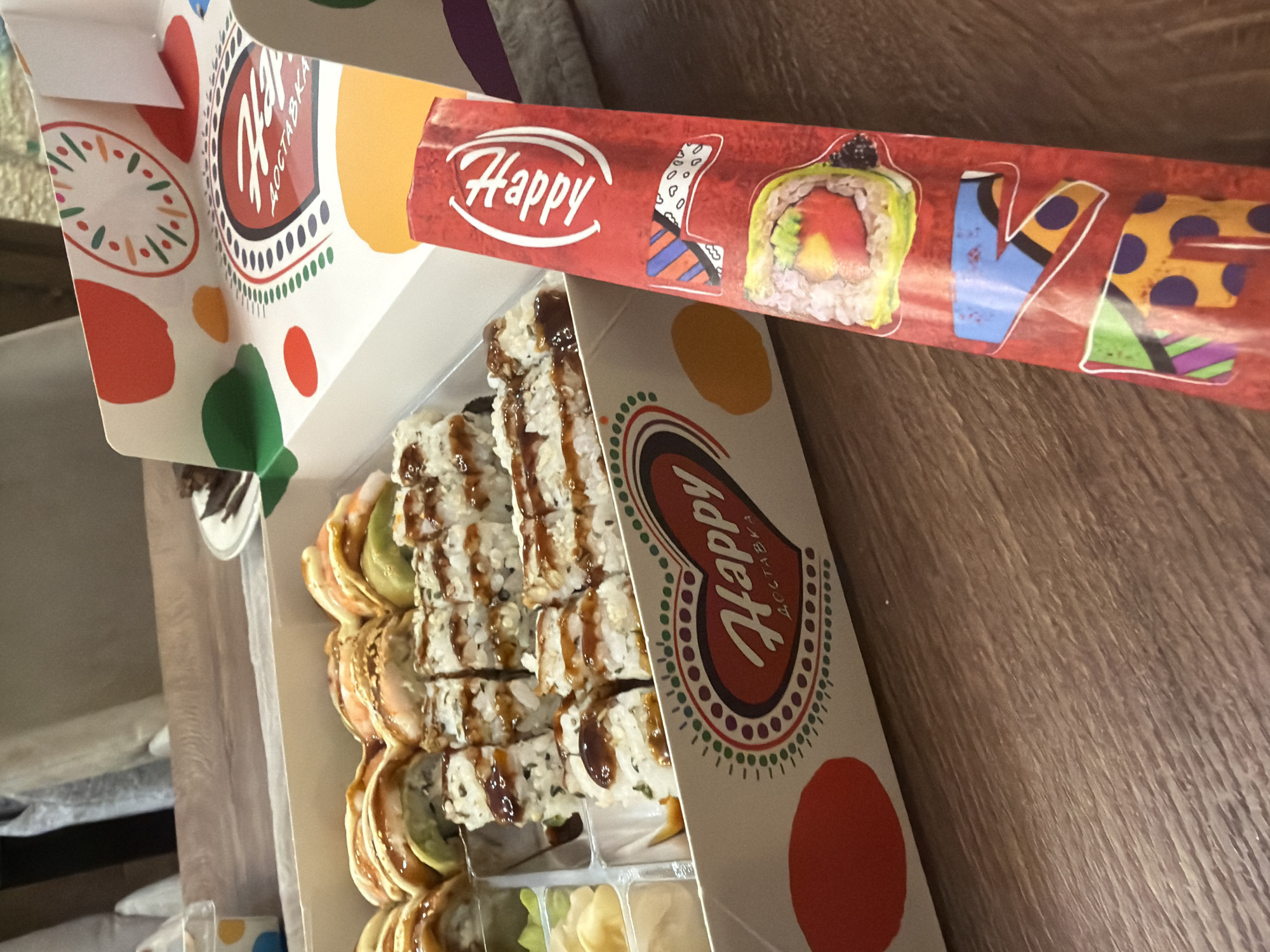 A close-up, high-angle shot shows a white cardboard sushi box filled with various types of sushi and rolls, placed on a wooden table. The box has a colorful, abstract design with red, orange, green, and dotted circles. A red wrapper with the word "Happy" in white font and some colorful graphic elements is partially visible in the foreground, leaning against the sushi box. The background is blurred, showing hints of furniture and possibly another box. The lighting is natural, coming from the left, creating soft shadows. No people are visible in the image.