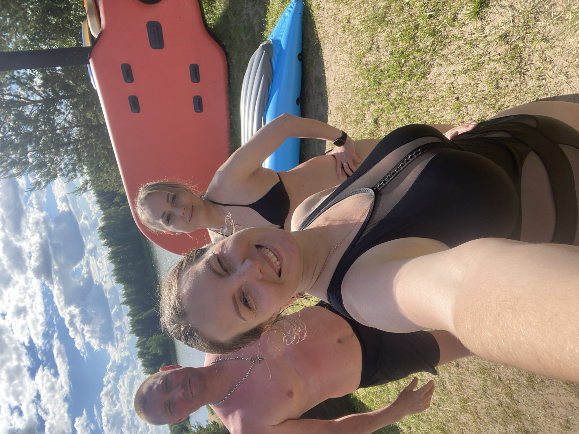 In Novyi, Russia, a selfie is taken on a sunny day at a waterfront location, possibly a beach or lake. Three individuals are present. In the foreground, a woman with long brown hair smiles at the camera, wearing a black swimsuit with mesh detailing. To her left, a man with a shaved head and a silver chain stands shirtless, wearing black shorts. Behind the woman in the foreground and to the right, another woman with blonde hair tied back wears a black bikini. In the background, a red inflatable raft and a blue inflatable kayak are visible on the sandy ground. The scene is bordered by a body of water and a dense line of green trees under a bright blue sky with scattered clouds. The overall atmosphere suggests a recreational outing.