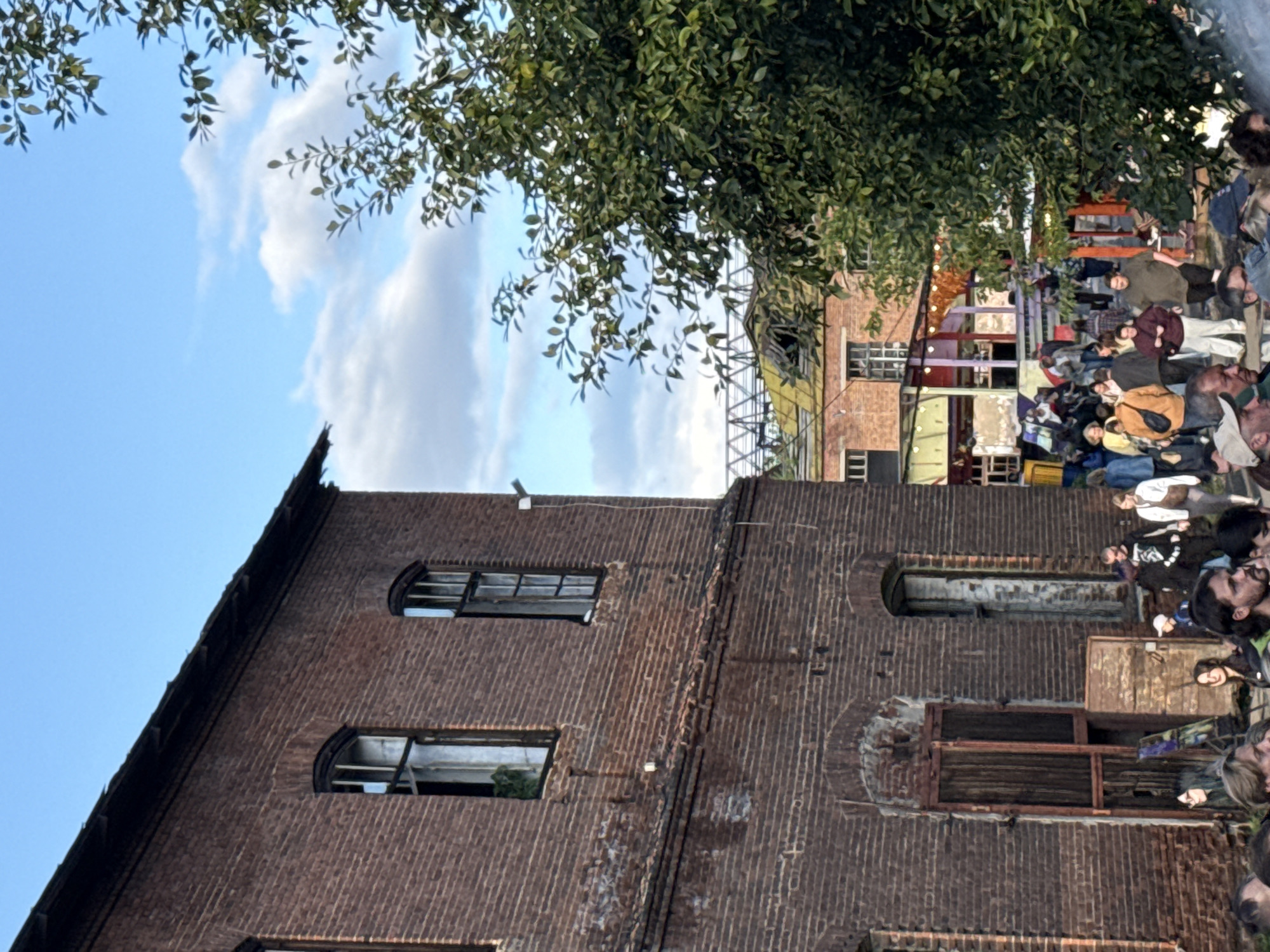 In Sysert, Russia, a weathered brick building stands next to a vibrant gathering of people. The building, with its arched windows and distressed wooden doors, suggests an industrial past. Sunlight illuminates the scene, casting shadows and highlighting the textured brickwork. A lush green tree with dense foliage on the right provides a natural contrast to the urban architecture. In the background, more buildings and structures are visible, some appearing to be under renovation or having a colorful, festive appearance. A crowd of diverse individuals are interacting in the space between the buildings, suggesting a social event or community gathering. The sky above is a clear blue, dotted with scattered white clouds.