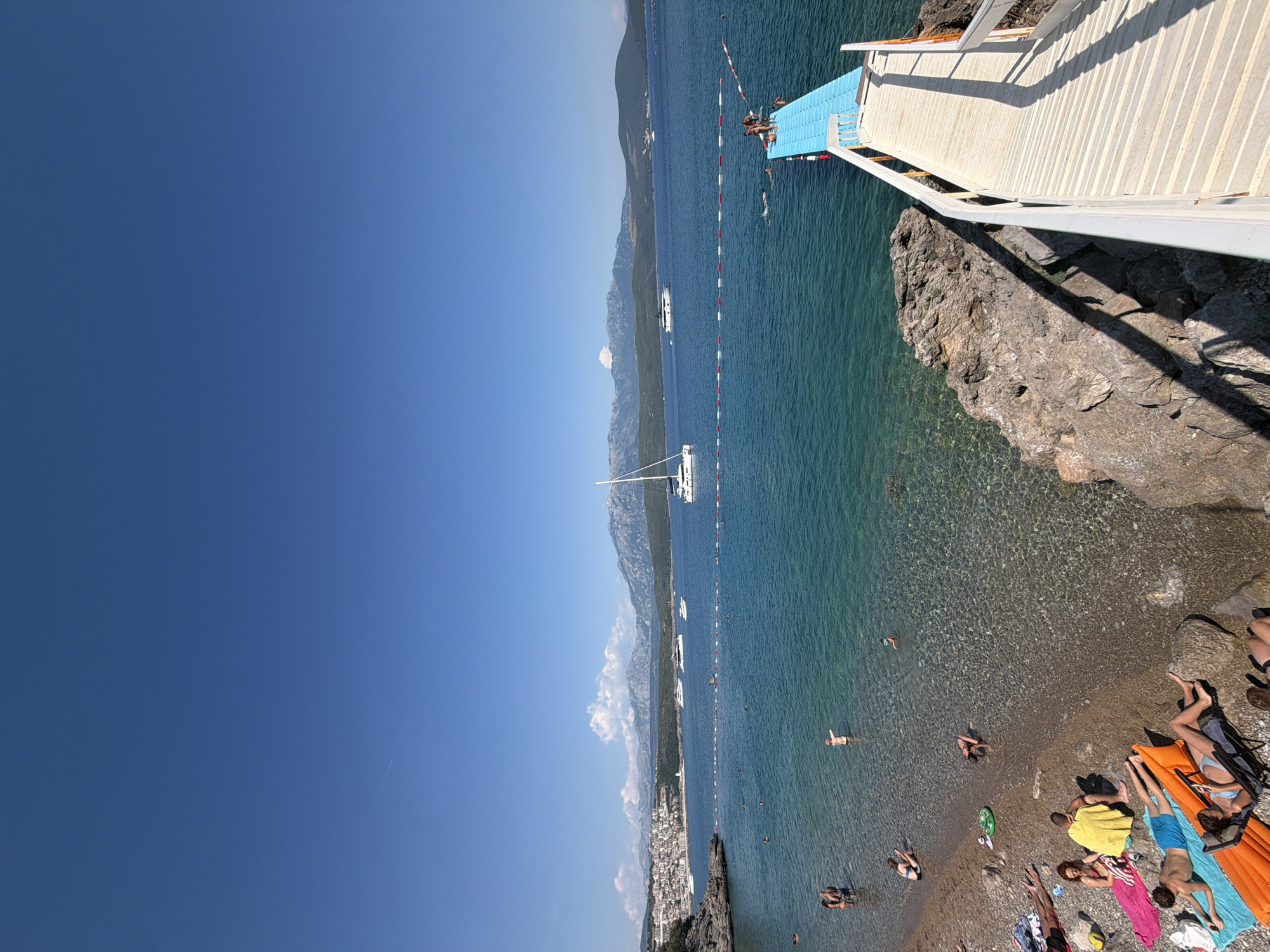 The image depicts a sun-drenched scene at a beach in Radovići, Montenegro. In the foreground, a wooden ramp with white railings leads down towards a bright blue floating platform extending into the clear, turquoise water. Several people are visible on the platform, some standing and others near the edge. The water is transparent, allowing the pebbled seabed to be seen, and a red and white rope barrier demarcates a swimming area in the sea. A small number of people are swimming or wading in the water.
The shoreline, composed of pebbles and rocks, is occupied by sunbathers. Various individuals are lying on towels and loungers, some partially clothed and others in swimwear. A few people are seated or standing on the beach.
Further out in the bay, several boats are anchored, including a white sailboat and two yachts. In the distance, a coastal town with buildings stretches along the shoreline, backed by a range of mountainous terrain under a clear blue sky. The overall atmosphere is one of a leisurely summer day at a coastal resort.