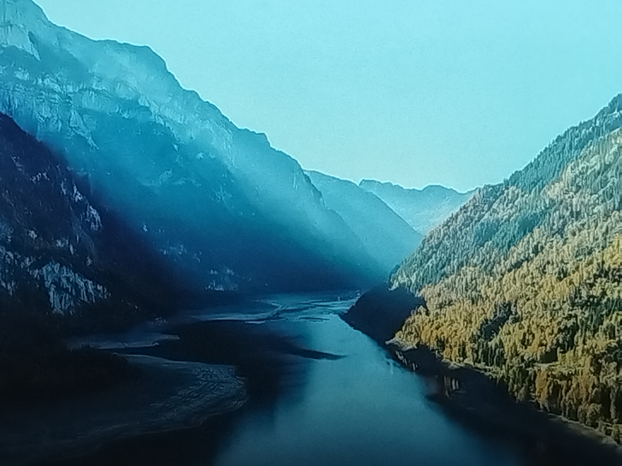 The image depicts a serene and majestic mountain landscape. Towering mountains flank a tranquil body of water, likely a river or a lake, that snakes through the valley. The slopes of the mountains on the left are rugged and shrouded in a cool blue mist, suggesting a possible mountainous region with sparse vegetation or rocky terrain. In contrast, the mountains on the right are densely covered with trees, displaying a mix of green and yellow hues, possibly indicating autumn foliage or a forest that thrives on that side of the valley.
The lighting suggests either early morning or late afternoon, with soft rays of light piercing through the atmospheric haze, creating dramatic shadows and highlighting the contours of the mountains. The sky is a clear, pale blue. The overall mood is peaceful and awe-inspiring, evoking a sense of natural grandeur and solitude. There is no visible text in the image. The provided location context of Balashikha, Russia, does not align with the typical mountainous terrain shown, suggesting this may be an error or that the image is meant to represent a general landscape.