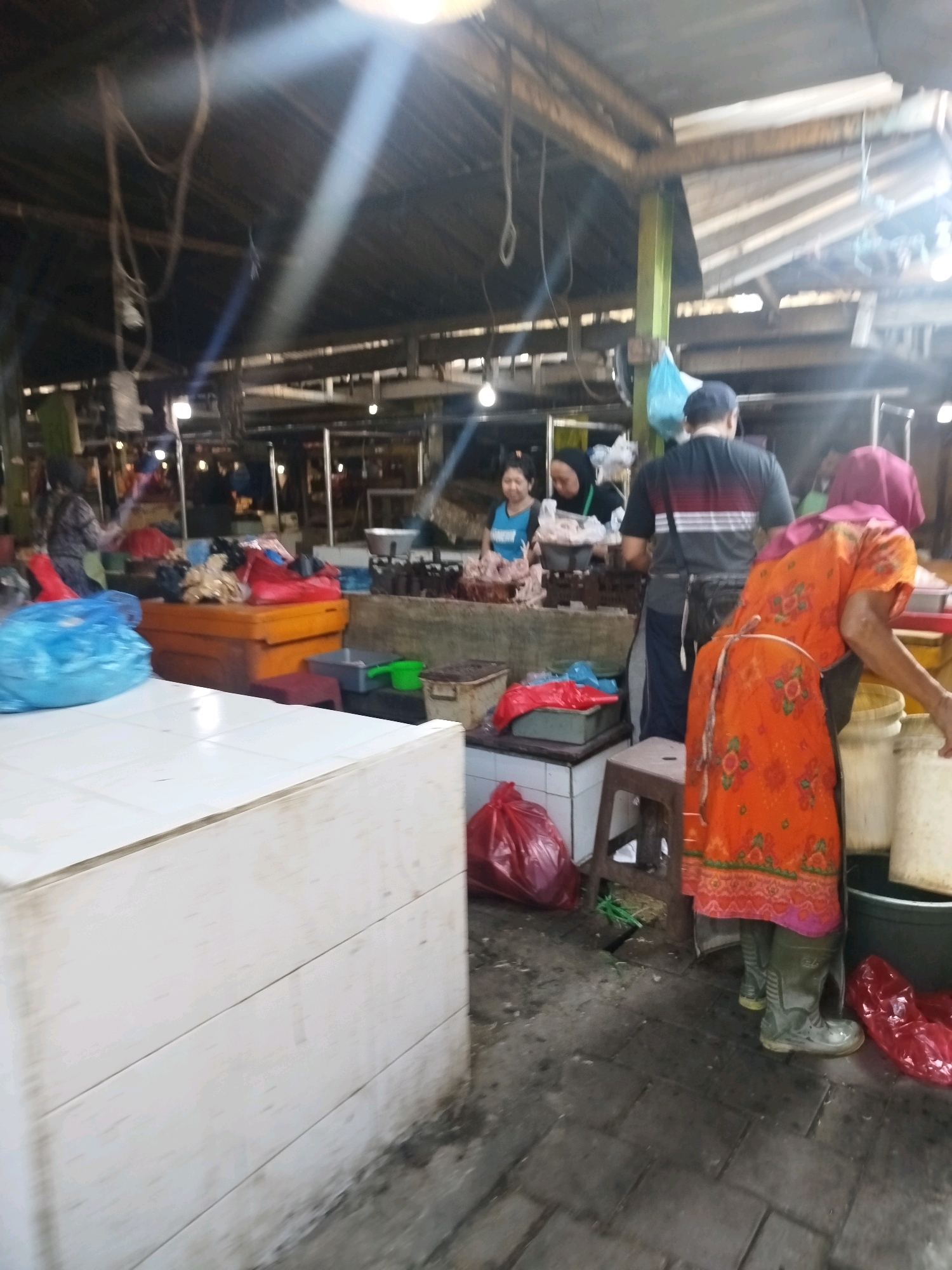 The image depicts a market scene in Larangan, Indonesia, likely a traditional market. The setting is an open-air or semi-covered structure with a visible wooden roof and exposed beams. In the foreground, a white tiled counter or stall is prominent. To the right, a person wearing an orange patterned apron and green boots is handling large white containers. Several red plastic bags are visible on the ground, along with some bundles of green produce. In the background, other market stalls and vendors are present. One vendor, wearing a blue top, appears to be serving customers. Another person in a black and red striped shirt and a blue cap is also visible. The overall atmosphere suggests a busy marketplace with various goods being displayed and sold, likely including fresh produce or meats given the context of a market. The lighting appears to be a mix of natural light filtering through the roof and artificial lights.