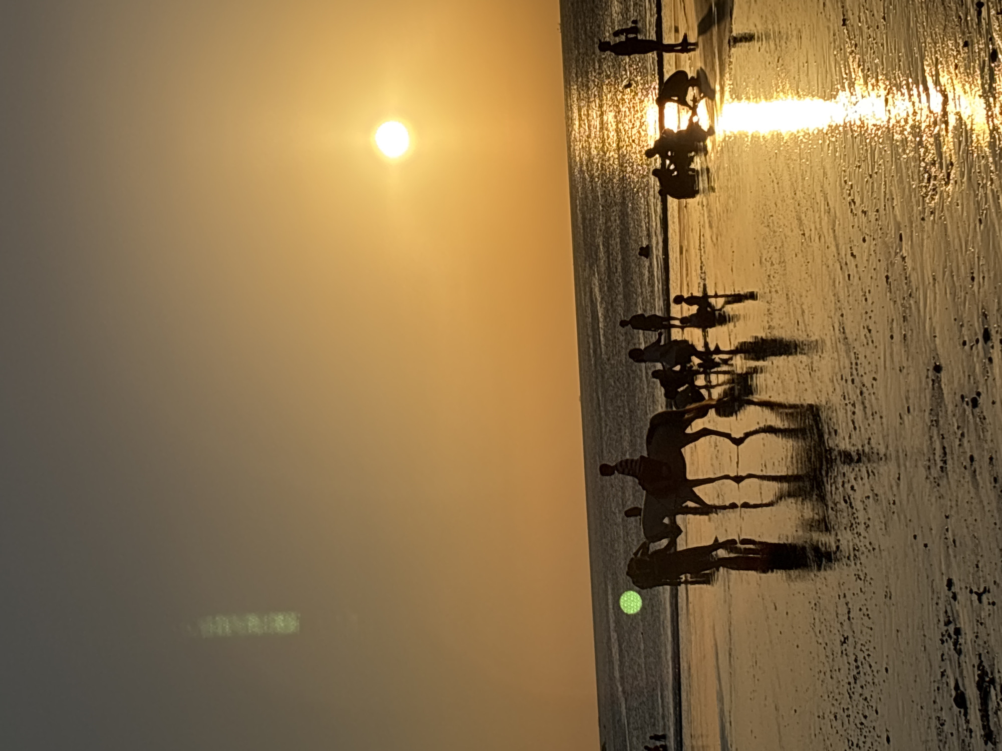 The image depicts a sunset scene at Kuta beach in Kuta Selatan, Indonesia. Silhouettes of people are visible on the sandy shore and in the shallow water. Several individuals are engaged in activities, including what appears to be horseback riding, with figures mounted on two horses in the mid-ground. Other groups of people are gathered, with some children playing near the water's edge. The sun, a bright orb, is setting over the ocean, casting a golden and orange glow across the sky and reflecting on the wet sand. The ocean is calm, with gentle waves lapping at the shore. The overall atmosphere is one of a relaxed evening at a popular beach destination.