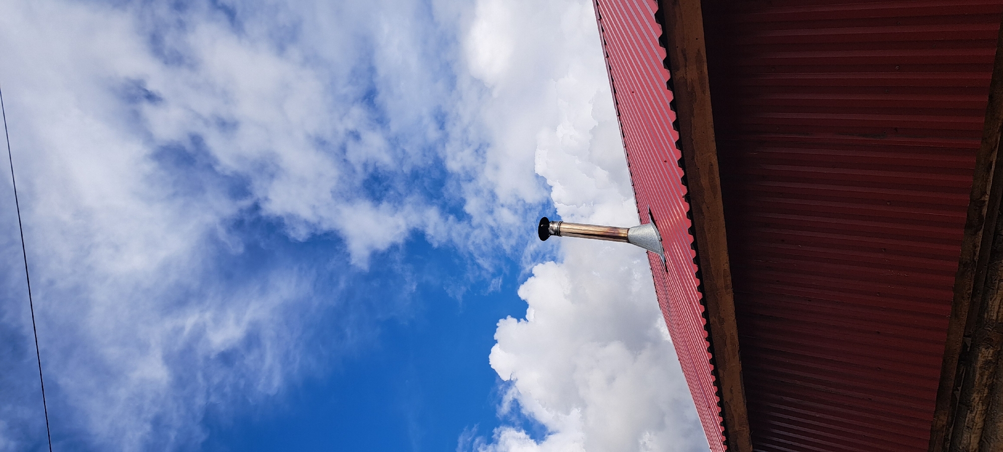A metal chimney protrudes from a red, corrugated metal roof of a building. The roof is angled, with a wooden beam visible at the edge. The sky above is blue with scattered white clouds. A thin black wire runs diagonally across the top portion of the image. The scene appears to be outdoors, possibly in a rural or suburban setting in Азьмушкино, Russia. There are no people or animals visible in the image.