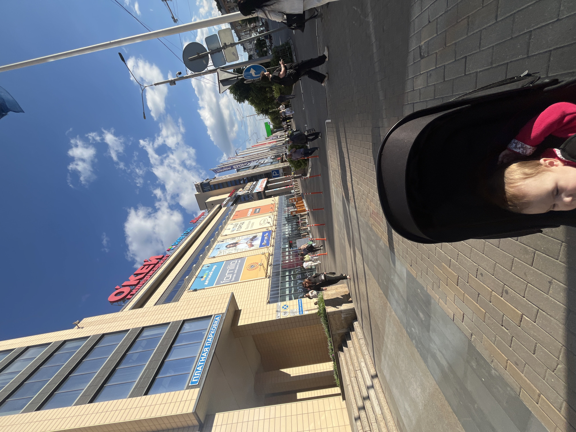 This image depicts a bright, sunny day in what appears to be a commercial area in Voronezh, Russia. The focal point is a large, modern shopping mall with a beige facade and dark-framed windows. Prominent signage on the building includes the word "ÖKEN" in large red letters and "ПЛАТНАЯ ПАРКОВКА" (Paid Parking) in blue. Other visible text on the mall's facade indicates "Детский мир" (Children's World) and "familia," suggesting a diverse retail environment.
In the foreground, a stroller is partially visible, with a baby inside wearing a pink outfit. The stroller is on a paved sidewalk made of brick-like stones. To the right of the stroller, the sidewalk continues, with a shaded area cast by an unseen structure.
Beyond the mall, a street is visible with traffic and pedestrians. Several people are walking or standing on the sidewalk in front of the mall. Overhead, utility lines and traffic signals are present against a clear blue sky with scattered white clouds. The overall scene suggests a typical day at a busy shopping center, with people engaged in shopping or leisurely activities. The bright sunlight and blue sky indicate it is daytime, likely late morning or afternoon. The presence of the stroller and the relaxed atmosphere of the people suggest a casual outing.