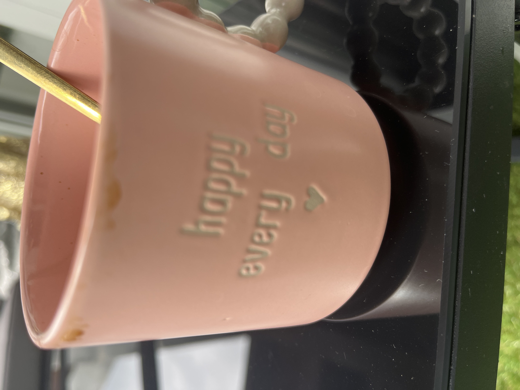 A close-up, eye-level shot focuses on a light pink ceramic mug with embossed text reading "happy every day" and a small heart. A gold spoon is visible inside the mug, partially submerged. The mug has a textured, beaded handle. There are a few light brown stains on the upper rim of the mug. The mug sits on a black, reflective surface. In the background, there is a blurred view of what appears to be a bright, possibly sunny environment with some green elements. The image does not provide any information about the city of Garmisch-Partenkirchen, Germany, nor does it depict any people.
