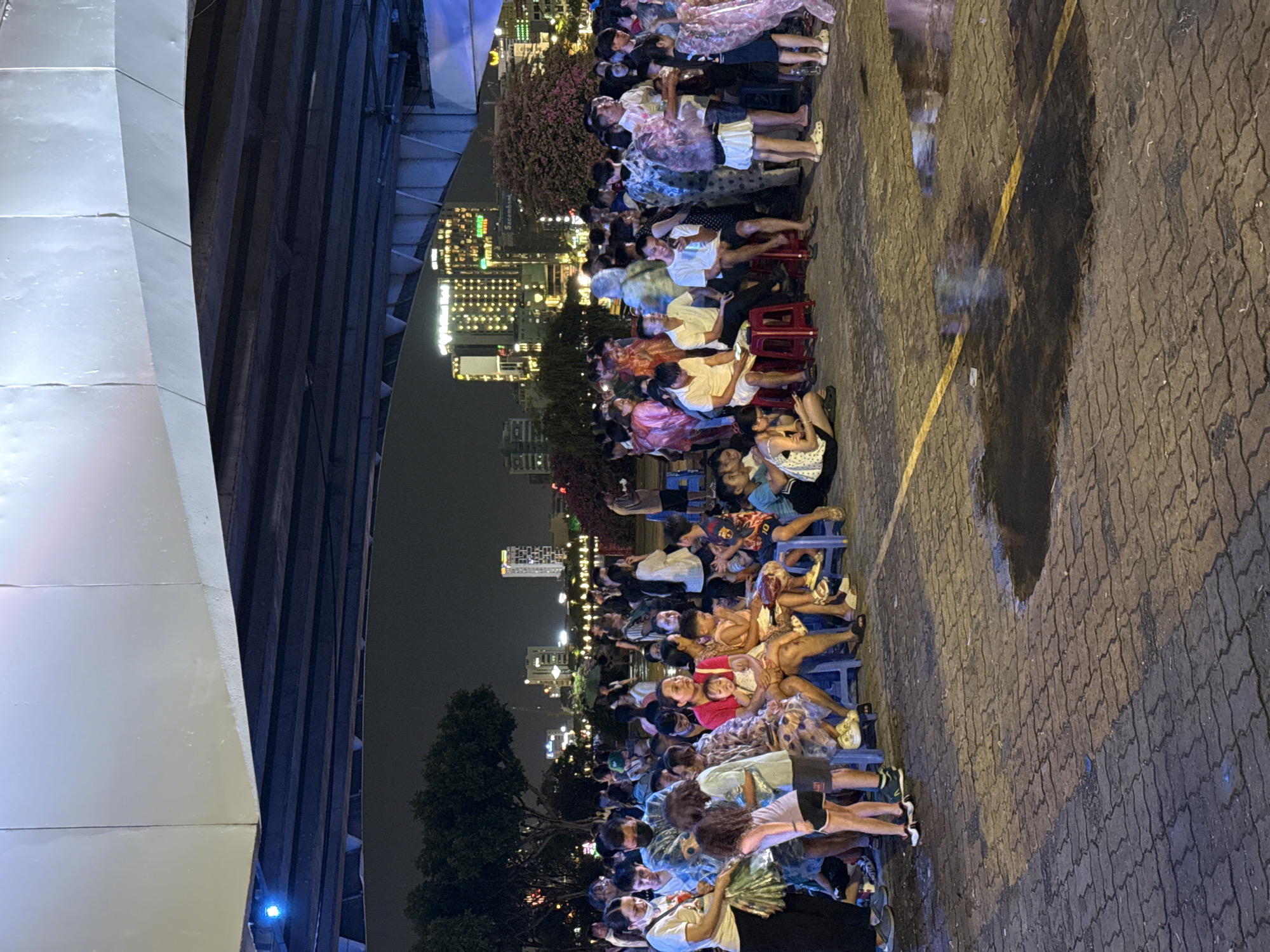 This is a nighttime scene under a bridge in Da Nang, Vietnam. A large crowd of people, predominantly Vietnamese, are gathered on a paved area, possibly for an event or performance. Many individuals are sitting on plastic chairs of various colors, including blue and red. Some people are standing and talking to each other, while others appear to be looking at something in front of them, out of frame. The atmosphere is festive, with some individuals wearing colorful clothing. In the background, the city skyline is visible with illuminated buildings and a body of water, suggesting a riverside or harbor location. The bridge overhead is lit with artificial lighting, casting shadows on the scene below. The paved ground shows signs of recent rain, with puddles reflecting the lights.