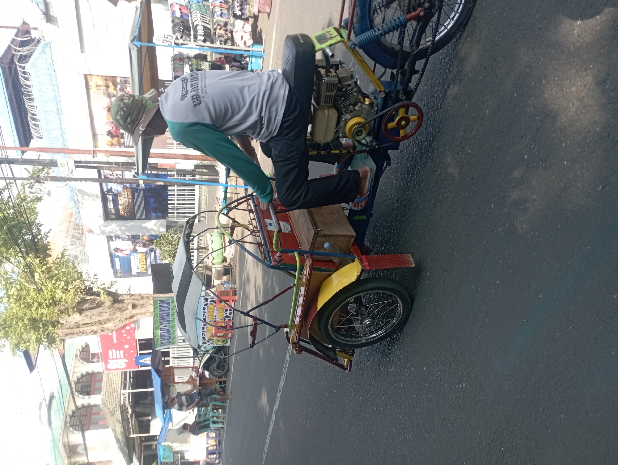 The image depicts a colorful, motorized tricycle, commonly known as a "becak," on a paved road in Nganjuk, Indonesia. The tricycle is equipped with a small engine and a covered passenger area, currently occupied by a person wearing a camouflage-patterned hat and a long-sleeved shirt with text that appears to be "UD. JATI INTI." The driver's legs are visible, and they are wearing blue flip-flops. The tricycle has a red, yellow, and blue color scheme, with various decorative elements. In the background, there are buildings, market stalls displaying merchandise such as shoes or sandals, and trees. The scene suggests a typical street environment in Nganjuk, likely a commercial or market area, with pedestrian and vehicle traffic.