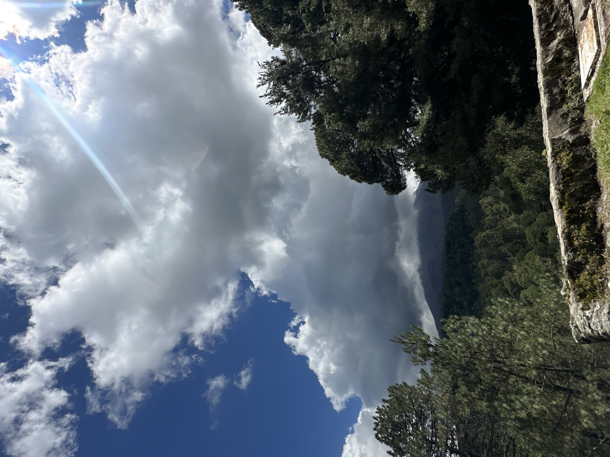 In San Mateo Ozolco, Mexico, this image captures a dramatic sky over a mountainous landscape. The sky is filled with a mix of bright white, fluffy cumulus clouds and darker, more ominous storm clouds, creating a stark contrast against the deep blue sky. Sunlight breaks through, casting a lens flare across the upper right portion of the image.
Dominating the foreground are tall evergreen trees, their dark green foliage casting shadows. In the middle ground, a dense forest stretches across rolling hills, leading the eye towards the distant mountain peak. The mountain itself is partially obscured by clouds and appears to be dormant, with its dark, rugged silhouette visible.
The foreground also features a low, stone wall or retaining structure, partially covered in moss and vegetation, suggesting a park or scenic viewpoint. The overall atmosphere is one of natural grandeur, with the dynamic weather patterns adding a sense of awe and perhaps a hint of foreboding. No people or animals are visible in the frame.