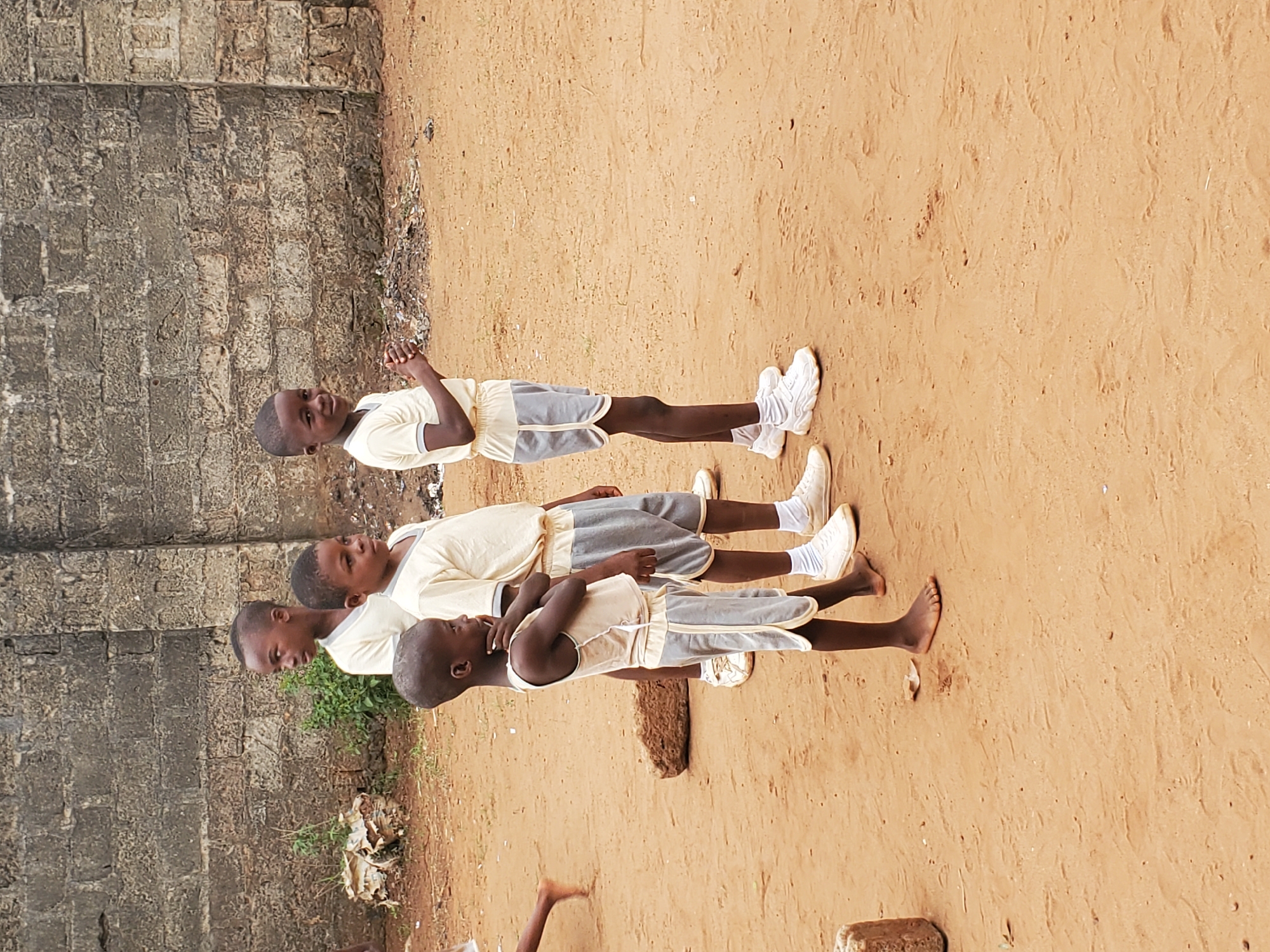 Four young boys are standing in a line on a sandy, reddish-brown surface in Ukwu-Abwa, Nigeria. They are wearing matching light-colored tank tops and grey shorts with a light-colored trim. Three of the boys are wearing white sneakers and white socks, while the last boy is barefoot. A grey concrete block wall is behind them, with some grass and debris growing in front of it.
