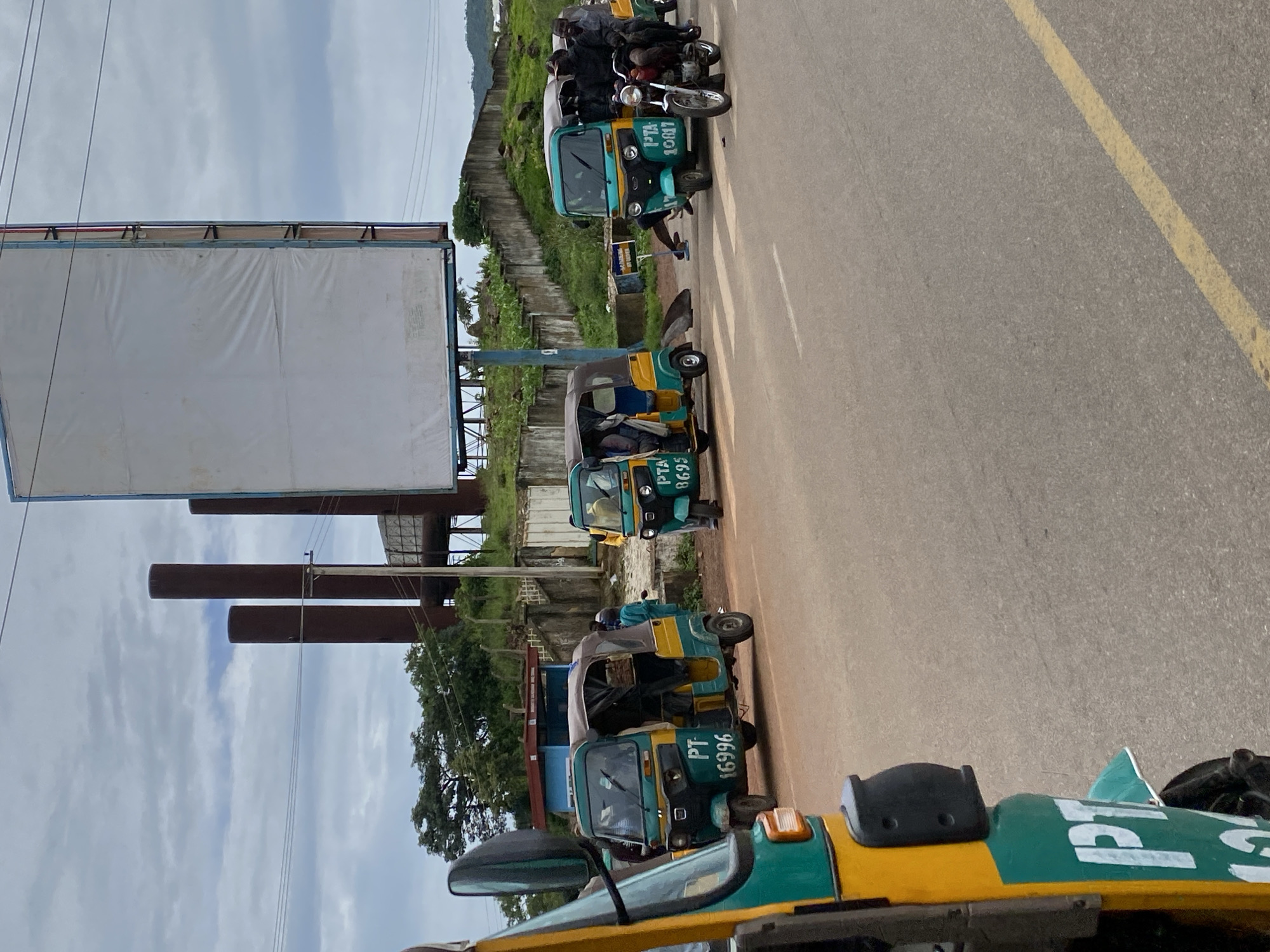 The image depicts a street scene in Rafin Par, Nigeria, featuring several auto-rickshaws, locally known as Keke Napep. The vehicles are primarily green and yellow. One auto-rickshaw in the foreground, partially visible, has "PT" and a partial number "12" in white lettering. Further down the road, another auto-rickshaw, also green and yellow, displays "PT 16996". To its right, a similar auto-rickshaw has "PTA 8695" on its side, with its doors open and occupants visible. Another auto-rickshaw, slightly further back and on the right side of the road, is marked "PTA 10817". A motorcycle with two occupants, one of whom is driving, is positioned in front of the "PTA 10817" auto-rickshaw. The road surface is asphalt with yellow lane markings. In the background, industrial chimneys or smokestacks are visible, along with a large, white, empty billboard mounted on a blue structure. There are also some trees and buildings in the background, suggesting an urban or semi-urban environment. The sky is cloudy. The scene appears to capture a moment of traffic or a waiting area for these public transportation vehicles.