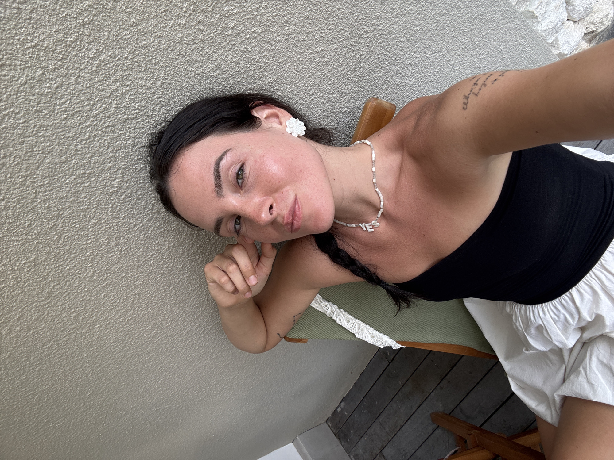 The image shows a woman sitting on a chair against a textured wall. She has dark hair styled in a braid and is wearing a black off-the-shoulder top and a white skirt. She is also wearing a beaded necklace with letter charms and white flower-shaped earrings. Her left arm is resting on the back of the chair, and she has a tattoo on her right arm. The background appears to be an outdoor setting with wooden flooring visible on the left. The scene is captured in a close-up, frontal view of the woman. The location is specified as Denpasar, Indonesia.