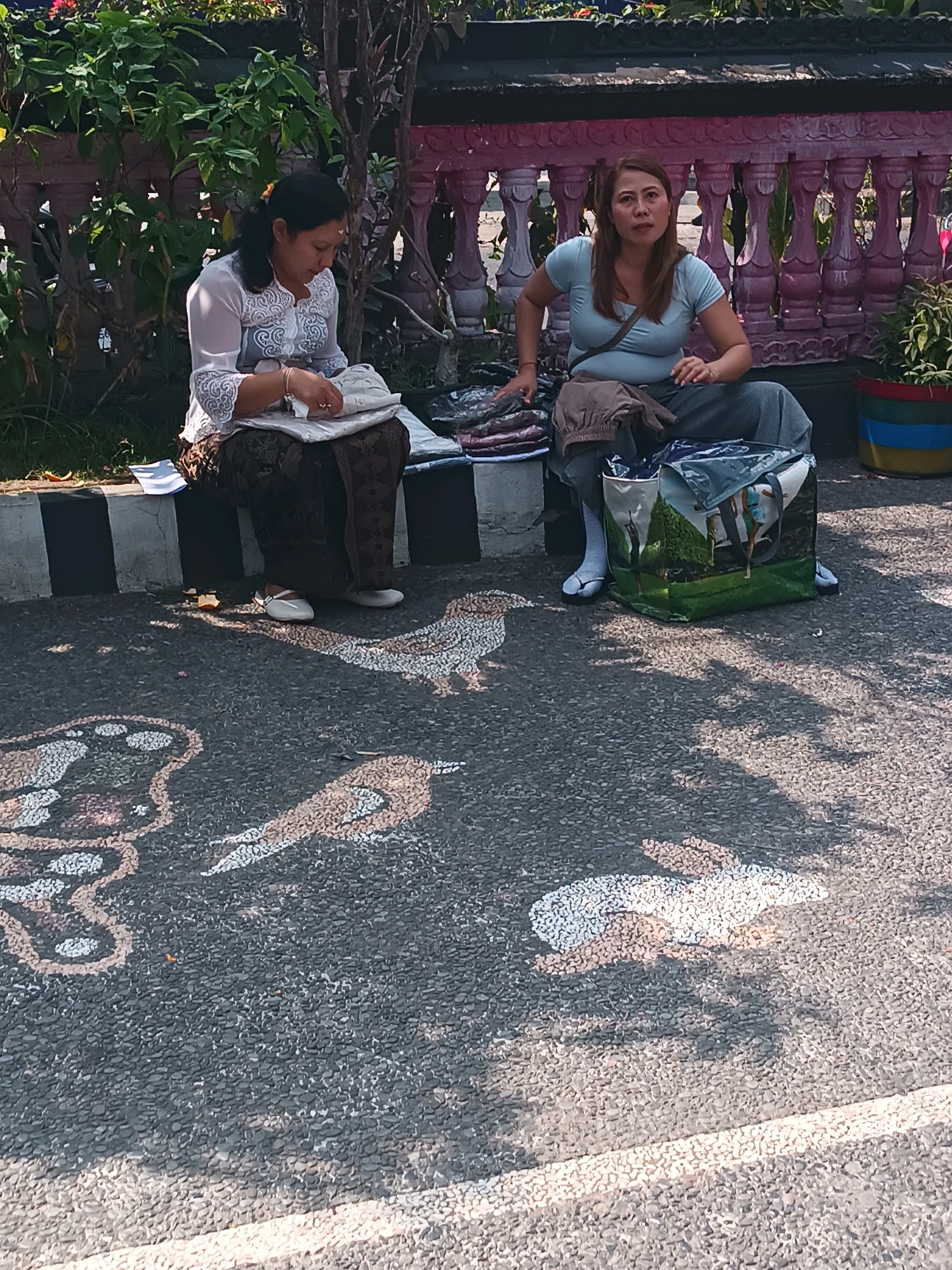 Two women are sitting on the curb next to a street with a decorative pattern painted on it, in Tabanan, Indonesia. The woman on the left is wearing a white semi-sheer blouse with intricate lace trim and a dark patterned sarong. She is holding a white package and appears to be sorting through it. The woman on the right is wearing a light blue t-shirt and grey pants. She is looking to her left with an open mouth and has a dark-colored bag with a picture of people on a grassy hill printed on it sitting beside her. The women are sitting in front of a pink decorative fence and lush green foliage. The street is paved with asphalt and has a black and white striped curb. The decorative pattern on the street appears to be made of small, light-colored pebbles embedded in the asphalt, depicting various animal shapes such as birds and rabbits.