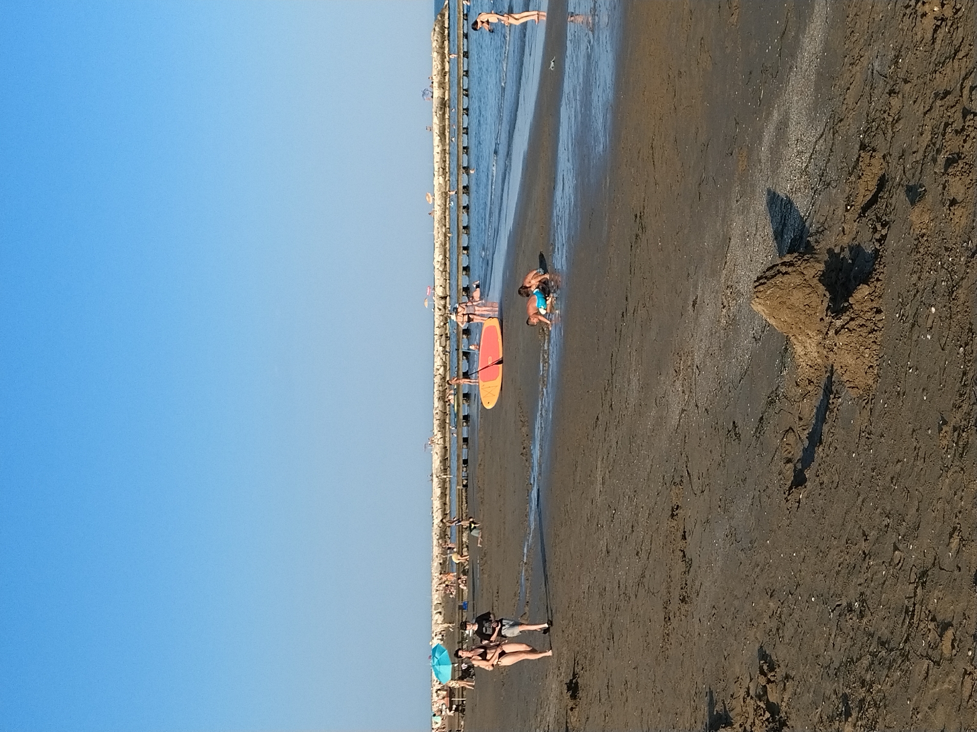 The image depicts a crowded beach scene in Caorle, Italy, under a clear blue sky. In the foreground, the wet sand is marked with footprints and tire tracks. A pile of sand sits to the right of center.
Further back, two individuals are walking on the sand, one in a bikini and the other in shorts and a t-shirt. A blue umbrella stands nearby. In the shallow water, two young boys are playing in the sand, one wearing blue shorts. A bright orange and yellow paddleboard is partially submerged near them.
The background shows a breakwater structure extending into the sea, with many people on and around it. The water is calm, with some people swimming and others standing. The overall impression is of a busy summer day at the beach.