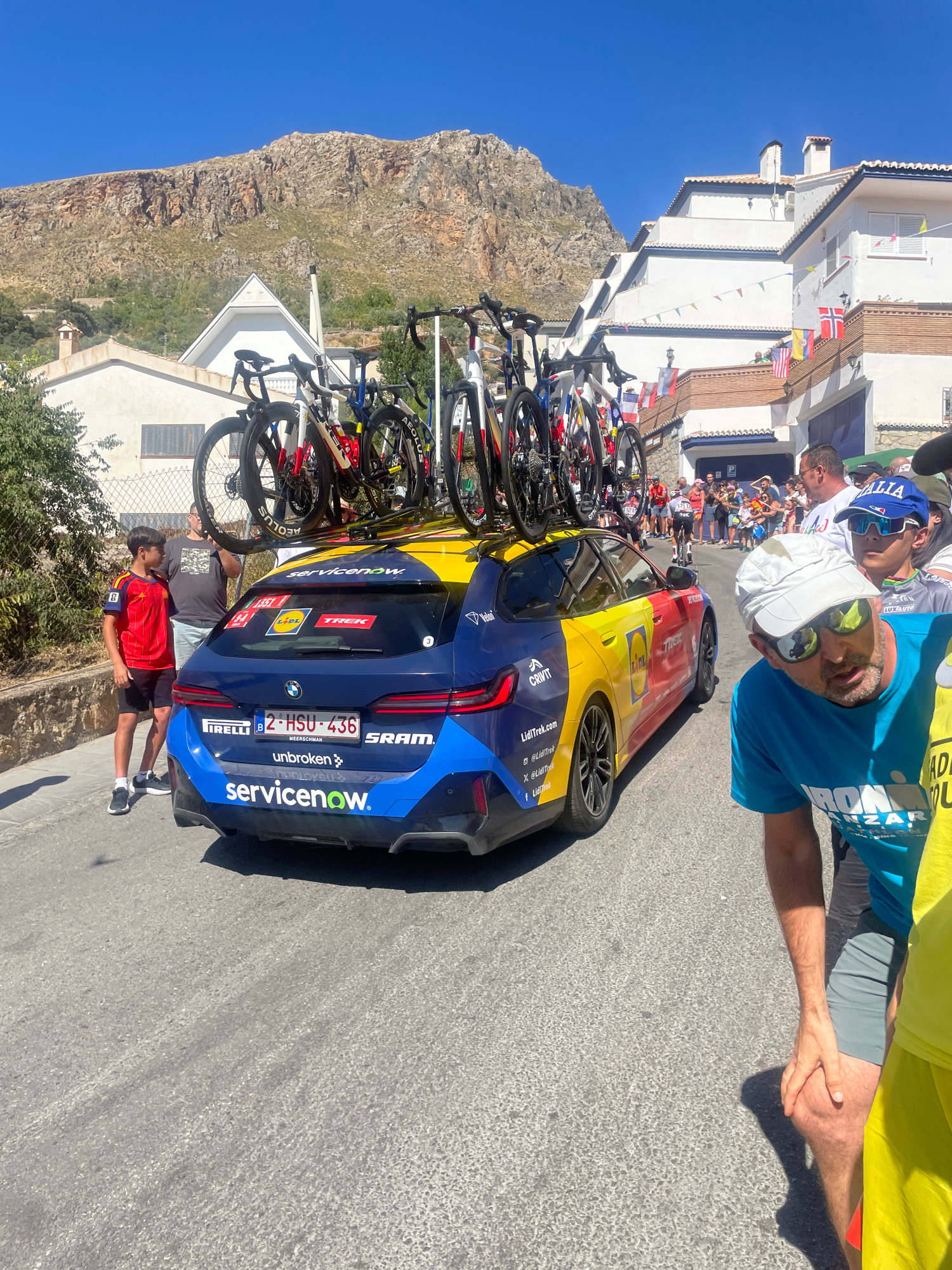 The image depicts a street scene in Güéjar Sierra, Spain, under a clear blue sky. In the foreground, a dark blue BMW wagon with a yellow and red design is parked on a paved road. The car is adorned with various sponsor logos, including "servicenow," "Pirelli," "SRAM," and "TREK." A rack on the roof carries multiple road bicycles. To the left of the car, a young boy wearing a Spanish national team cycling jersey and black shorts stands looking towards the car. Behind him, a man in a gray t-shirt and shorts is also visible. In the mid-ground, a group of spectators stands on a tiered walkway lined with colorful flags. Further back, more people are gathered, and a cyclist is visible on the road. The background features a steep, rocky mountainside and white buildings with red-tiled roofs. In the right foreground, a man in a blue t-shirt with "IRON..." visible on it, a white cap, and sunglasses is leaning down, appearing to be looking at the road. Another person in a bright yellow shirt is partially visible next to him.