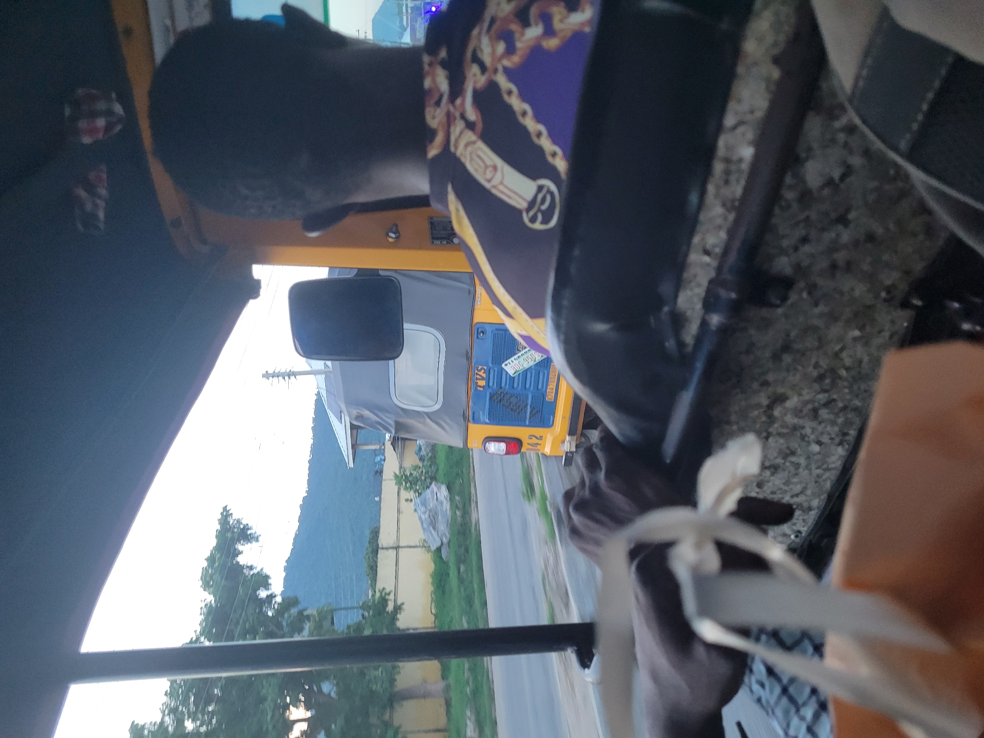 The media file shows a first-person perspective from inside a three-wheeled vehicle, likely an auto-rickshaw, in Lokoja, Nigeria. The view is angled, with the vehicle's interior partially visible. A person's head and shoulder are in the foreground, wearing a purple and gold patterned fabric. The steering wheel and part of the dashboard are also visible.
Outside the vehicle, another auto-rickshaw is visible, painted yellow and blue. It has the number "142" on the back and a license plate that appears to read "KOGI 955 CPG". The backdrop includes a road, green vegetation, and a hill covered in trees. The sky is bright, suggesting daytime, possibly late afternoon or early morning given the soft light. The scene appears to be taking place in an urban or semi-urban area. There is a sense of movement, as if the vehicle is traveling. The overall mood is casual and observational.