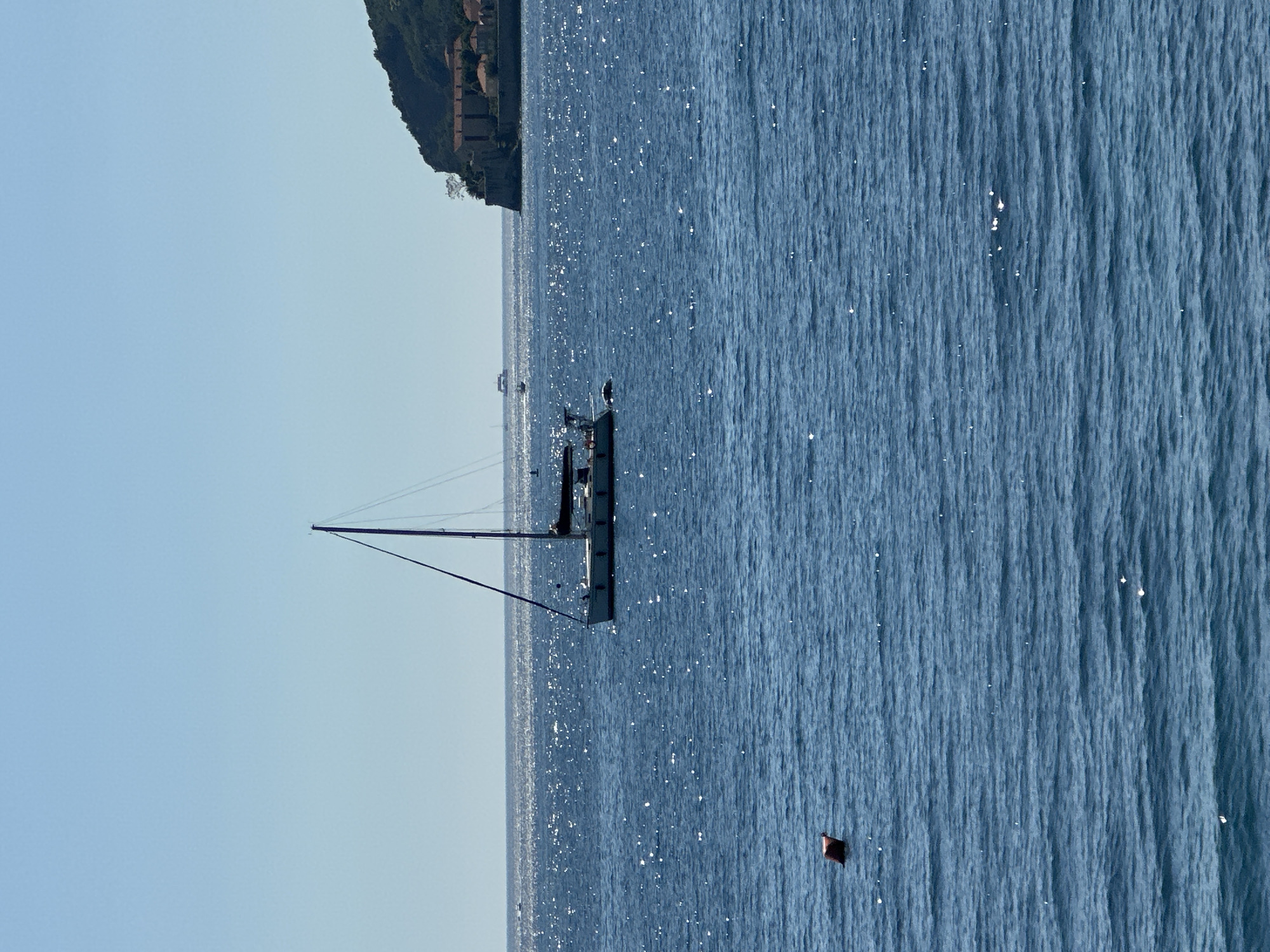 A horizontal outdoor shot shows a sailboat sailing on the Adriatic Sea on a sunny day near the city of Budva, Montenegro. The sailboat is off to the left side of the shot. The mast extends upward, and a line extends down to the left. A person is partially visible on the sailboat. A red marker is in the sea to the right of the sailboat. In the top left is a bit of land covered with trees and a few buildings.