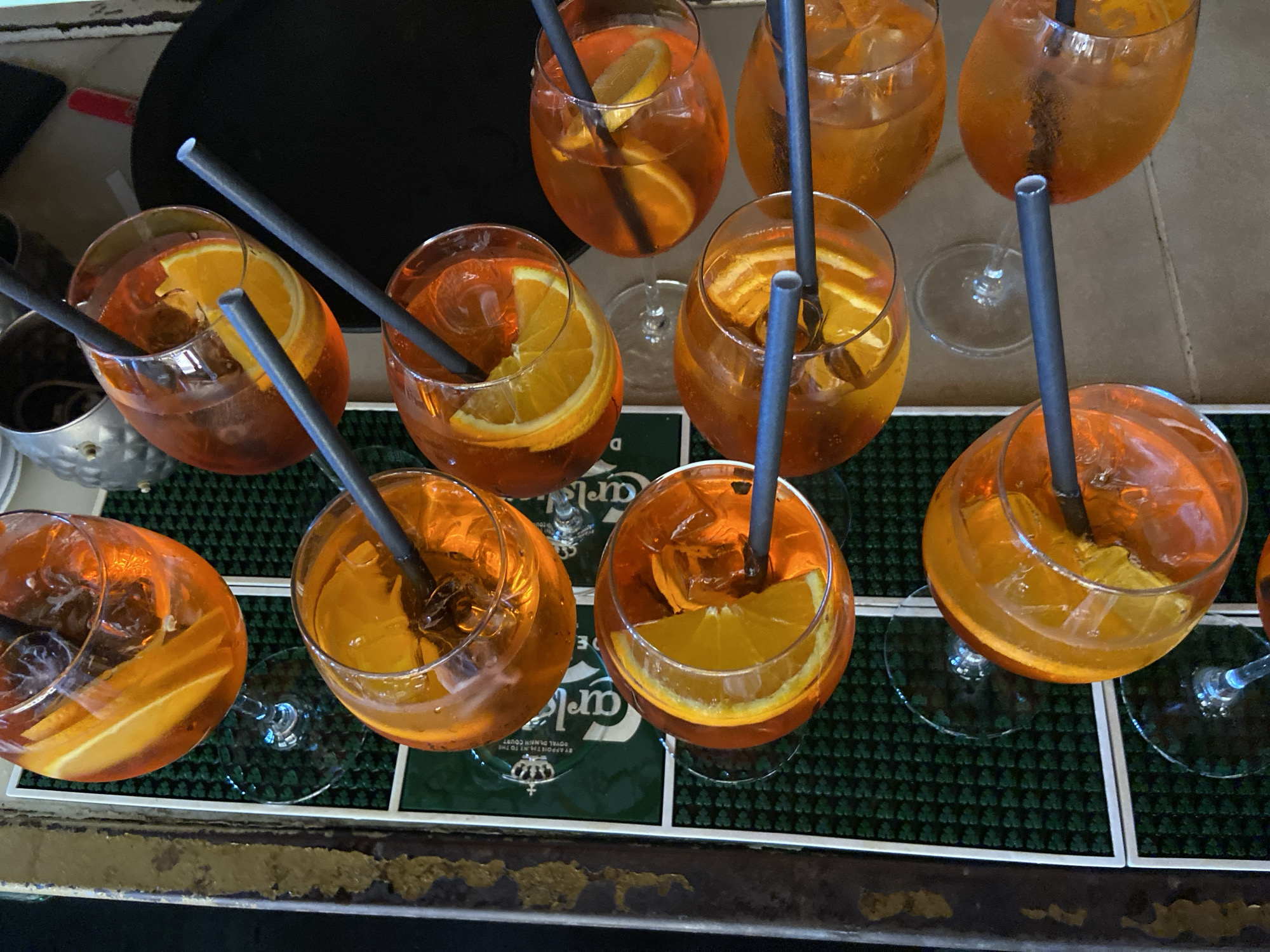 The image shows a collection of Aperol spritz cocktails arranged on a bar surface. There are nine cocktails visible, each served in a stemmed glass with ice, a slice of orange, and a gray straw. The cocktails are placed on a dark green, textured surface with white tiles that appear to be beverage mats or part of the bar top. One mat has a logo. The background is blurred, obscuring specific details but suggesting an indoor setting with other objects. The drinks are likely at a bar setting, possibly in Petrovac centar, Montenegro.