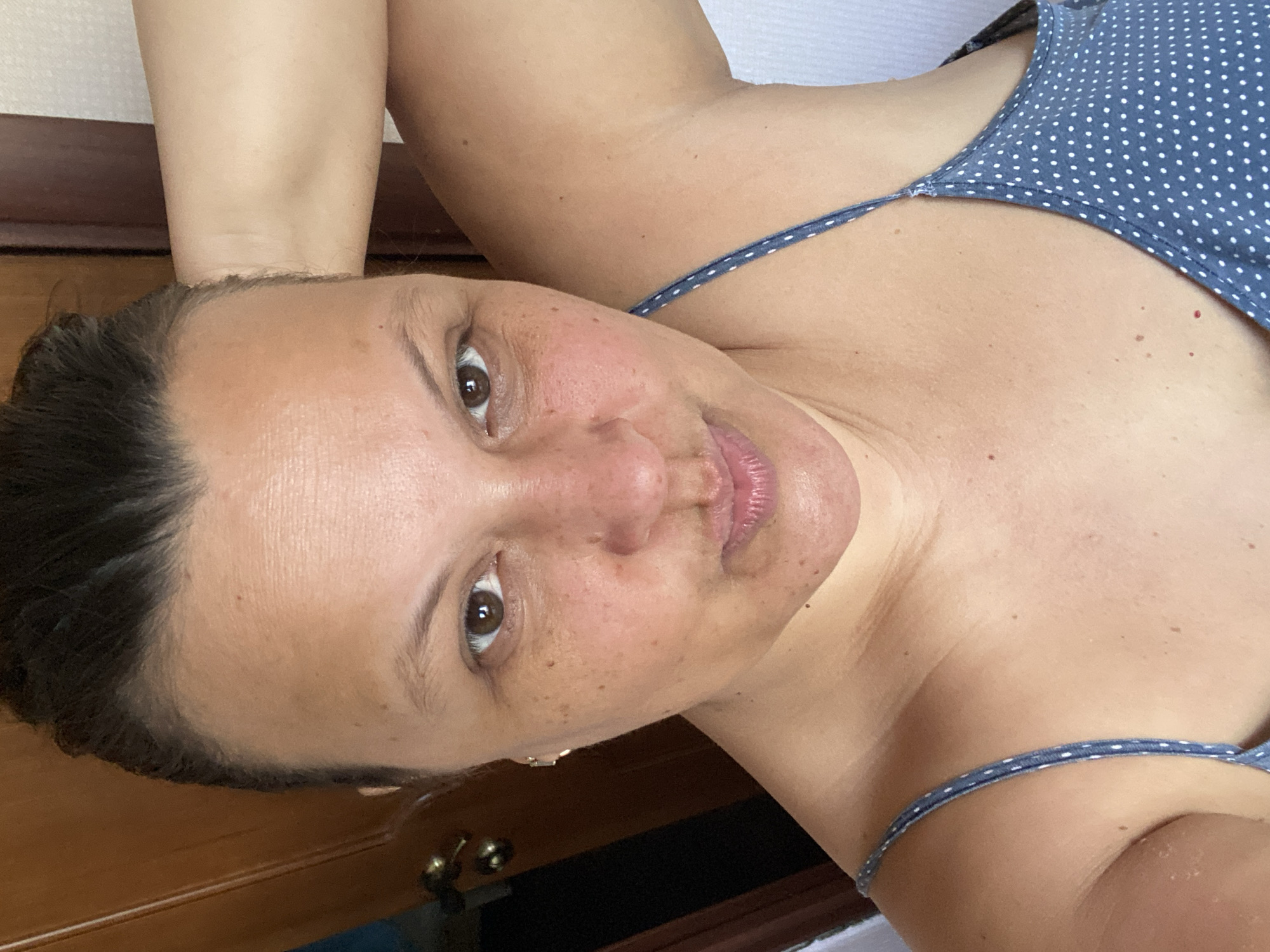 A close-up, tilted perspective photograph depicts a person with medium skin tone, brown eyes, and dark hair, wearing a blue and white polka-dot top with spaghetti straps. The individual's face is the primary focus, showing features like the nose, mouth, and freckles. The background includes a light-colored wall and a dark wooden surface, possibly a piece of furniture. The location is Unknown, Unknown.