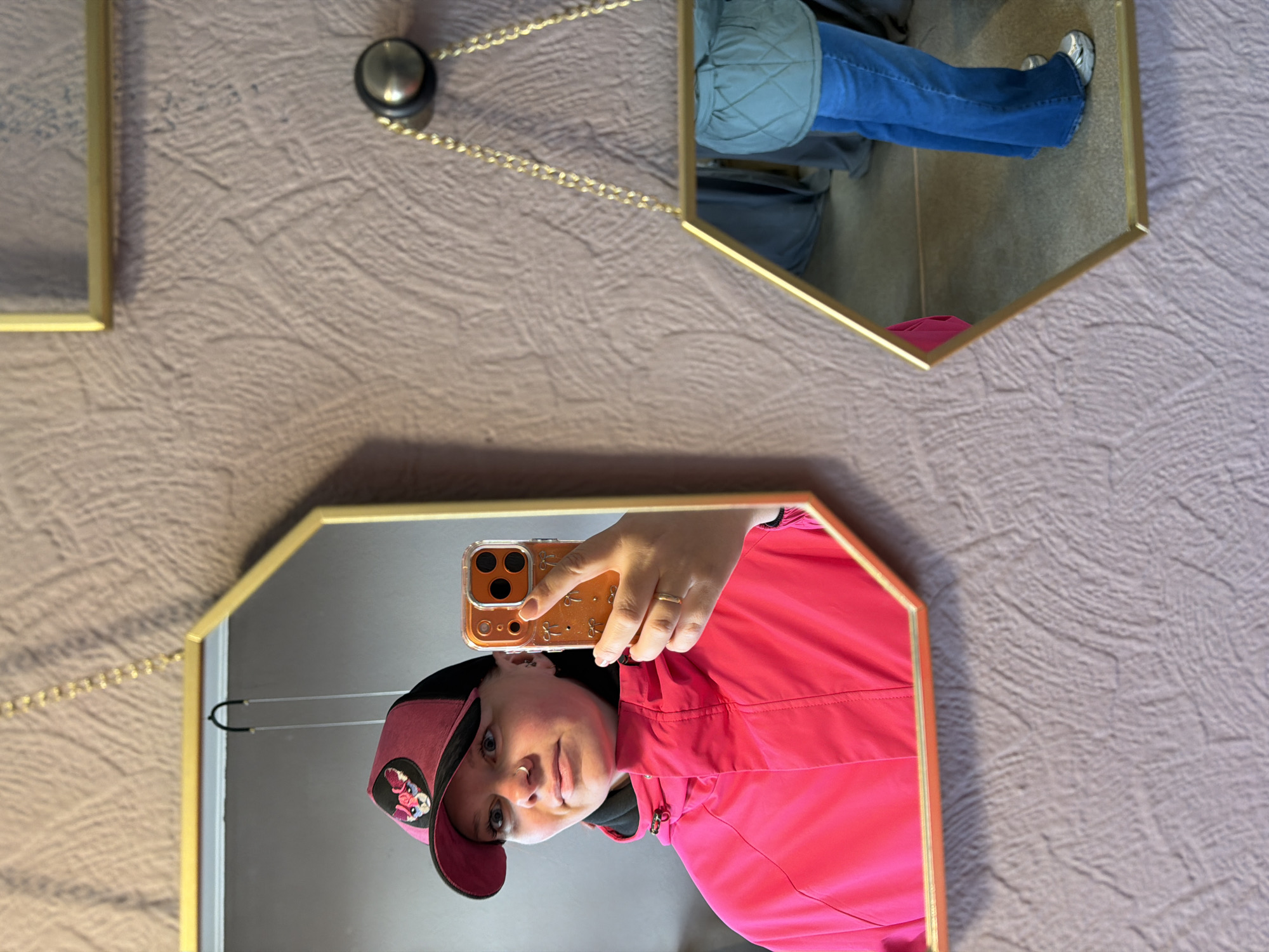 This image captures an indoor mirror selfie in what appears to be Chaikovsky, Russia. The scene features a light brownish-grey textured wall adorned with two gold-framed hexagonal mirrors, connected by a delicate gold chain.
In the larger, lower mirror, a person is visible from the chest up, taking the photo with a smartphone. They are wearing a vibrant hot pink jacket or windbreaker over a dark top. A two-tone cap, possibly black and pink with a colorful, indistinct patch or logo, covers their head. The person has light eyes and a silver nose ring on their right nostril. Their left hand, adorned with a simple gold ring, holds an orange or clear smartphone case featuring a repeating pattern, possibly small bunny ears. The individual looks directly into the phone, capturing their reflection.
The upper mirror reflects the lower body of a person, likely the same individual. They are wearing blue jeans and what appears to be the lower part of a light blue or grey quilted jacket or vest. Silver or white athletic shoes are visible on their feet.
The overall activity depicted is a casual mirror selfie, possibly taken in a home or a fitting room, with the mirrors serving as decorative elements on the wall. The lighting suggests daytime. No discernible text is visible on any objects or clothing.