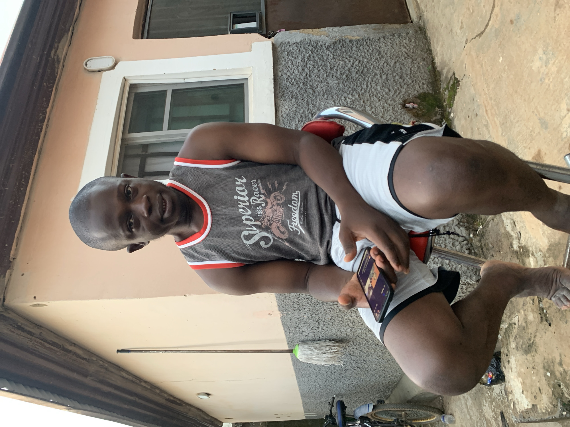 A dark-skinned man is sitting on a metal chair outdoors, holding a black smartphone. He is wearing a gray tank top with red trim and black and white shorts. The background features a light peach-colored building with a window and a gray wall. A mop and a bicycle are visible on the ground. The location is in Abuja, Nigeria.